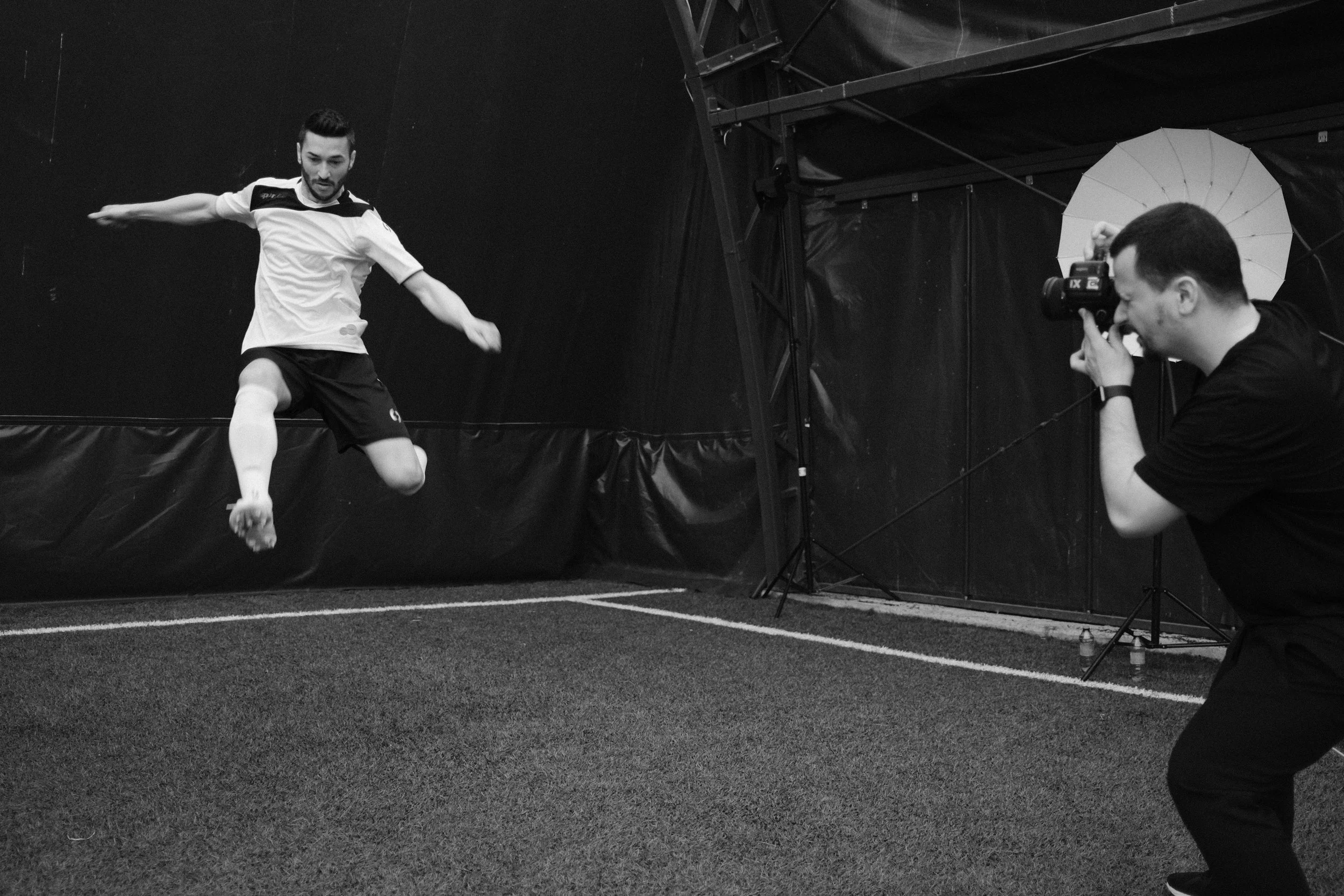 A man with a beard and mustache, wearing a sports shirt and shorts, is mid-air performing a jump or leap in an indoor sports facility. A photographer is taking photos of him, using a camera mounted with a flash, and is positioned on the right side of the image. The setting appears to be a sports arena or gym with dark walls and a cushioned floor.