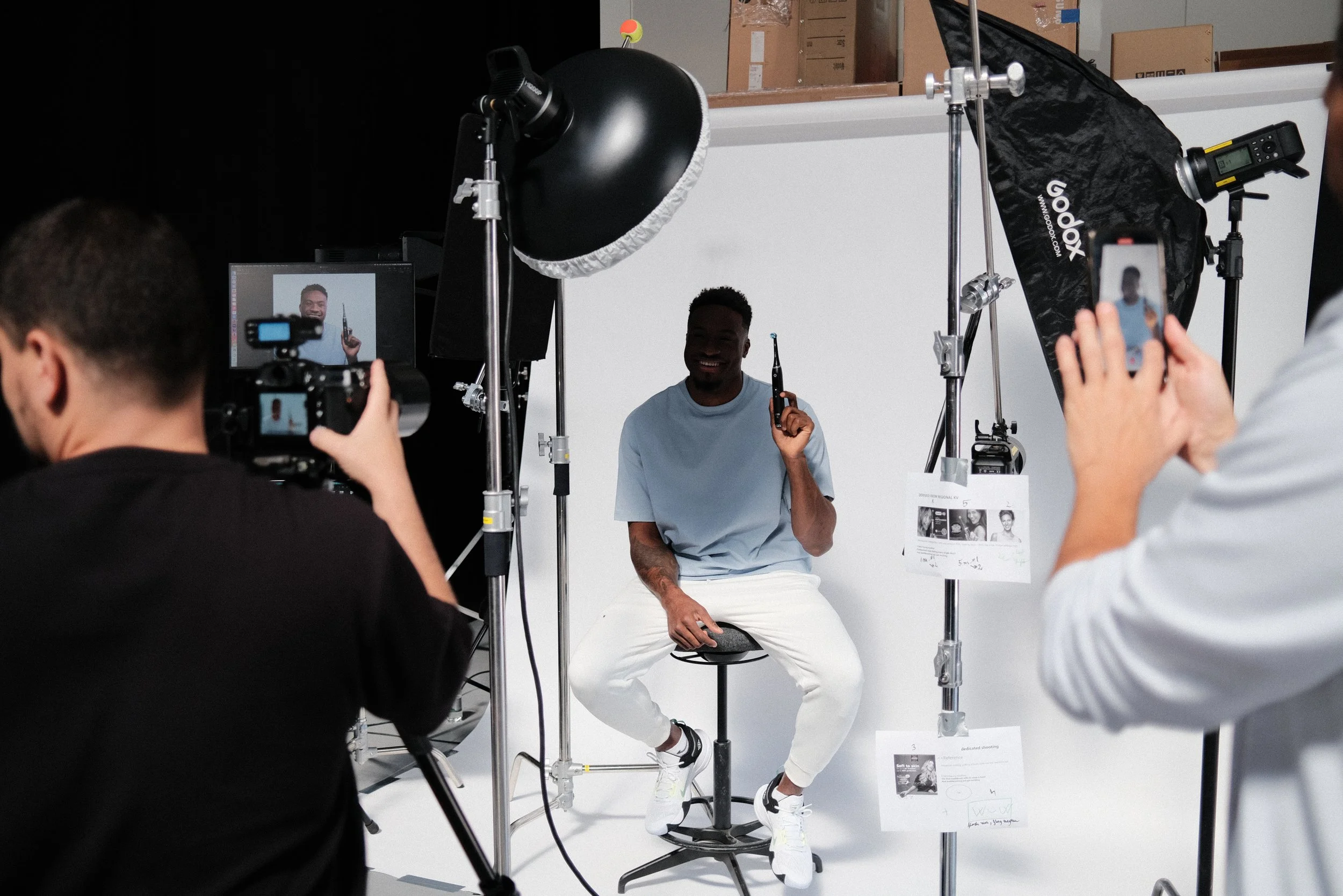 A photoshoot scene with a man sitting on a stool, smiling and holding a small object, surrounded by photography equipment including lights, cameras, and reflectors. Several people are taking photos and videos of him in a studio with a white background.