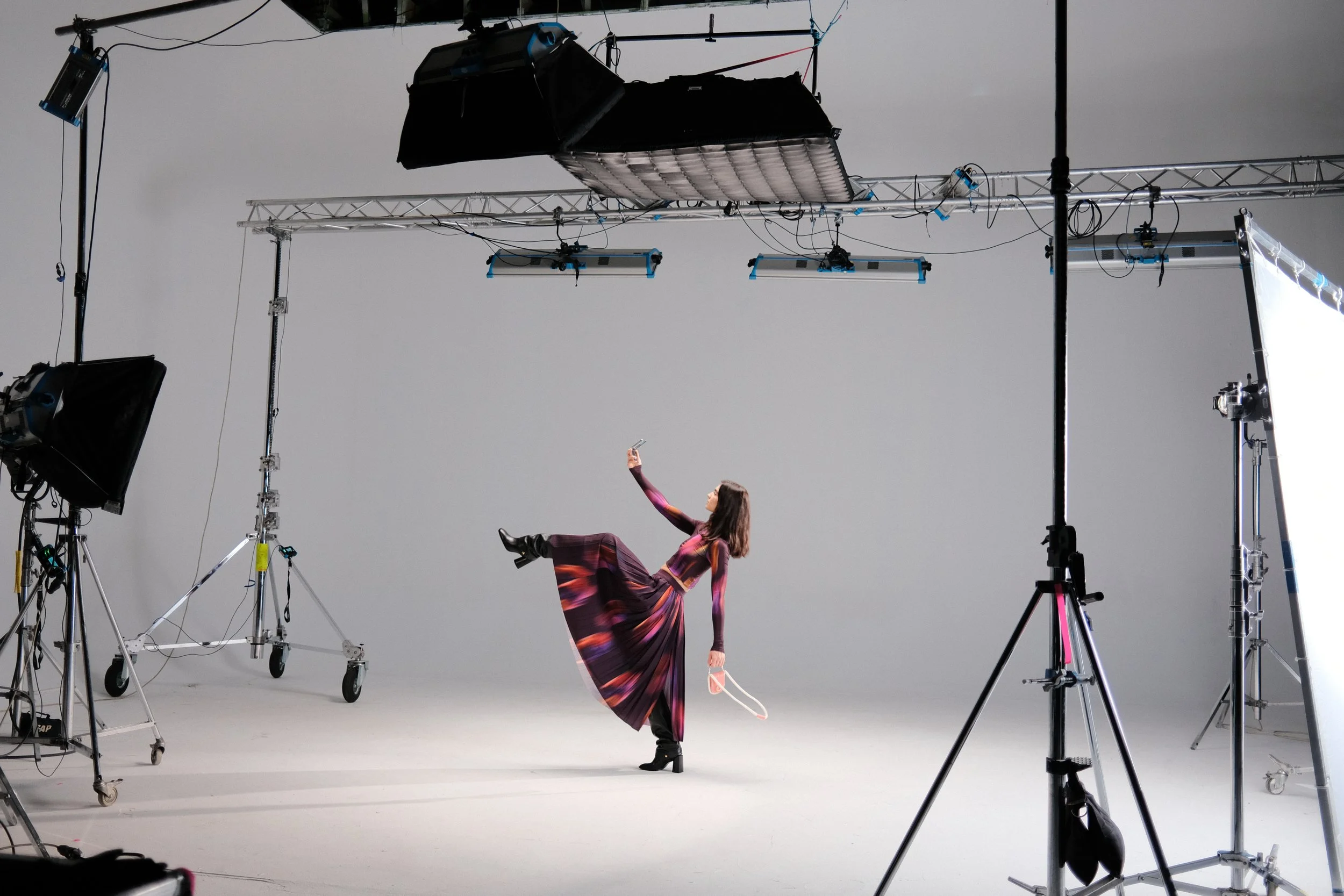 A woman in a colorful, flowing dress is lying back on her arm in a photo studio, holding a ring light in one hand and a pair of high heels in the other. The studio is filled with large professional lighting equipment and rigging.