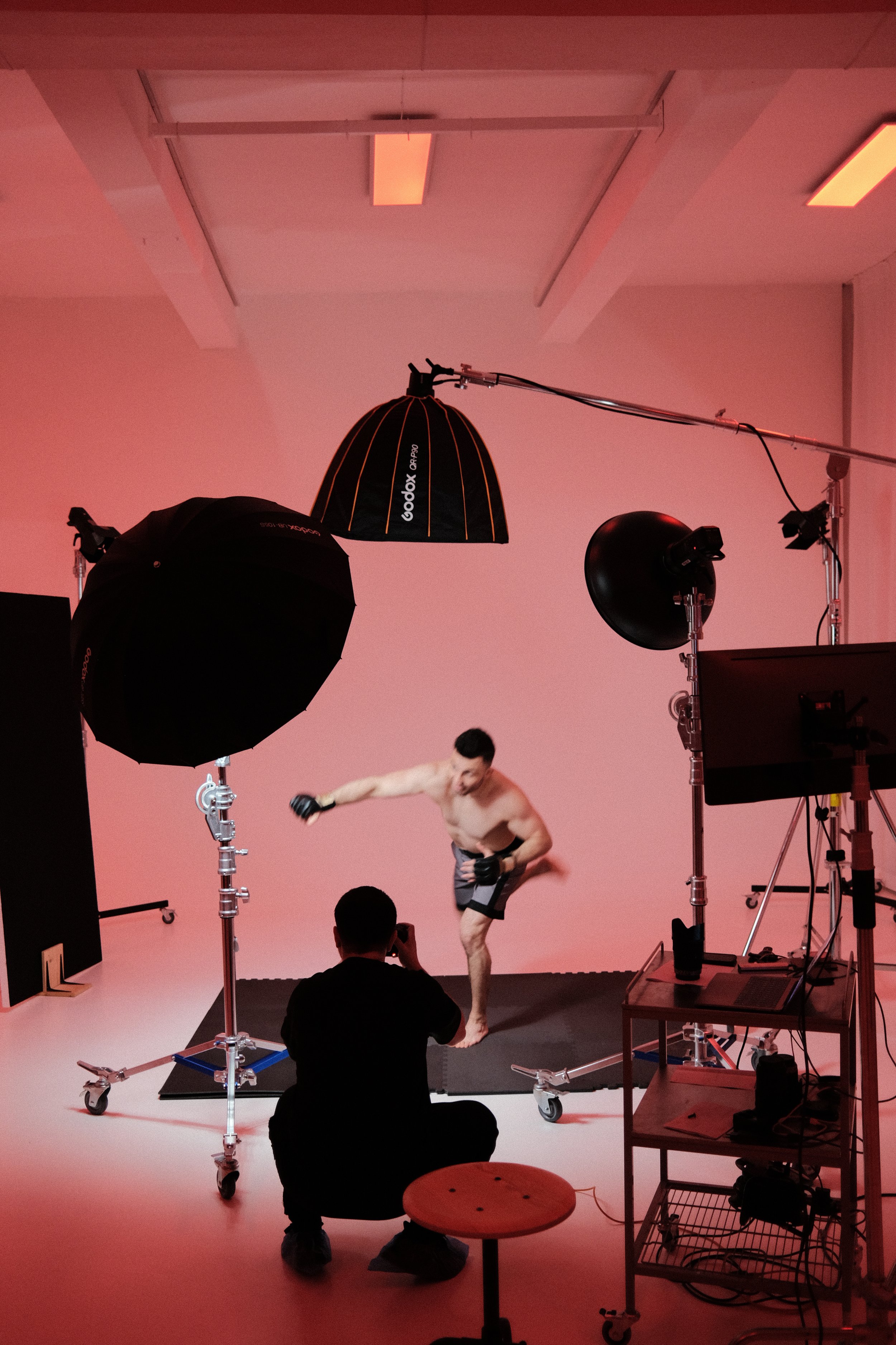 Studio photoshoot setup with lighting equipment and a model in athletic shorts and gloves practicing a punching motion. A photographer and an assistant are present, and a pink backdrop is visible.