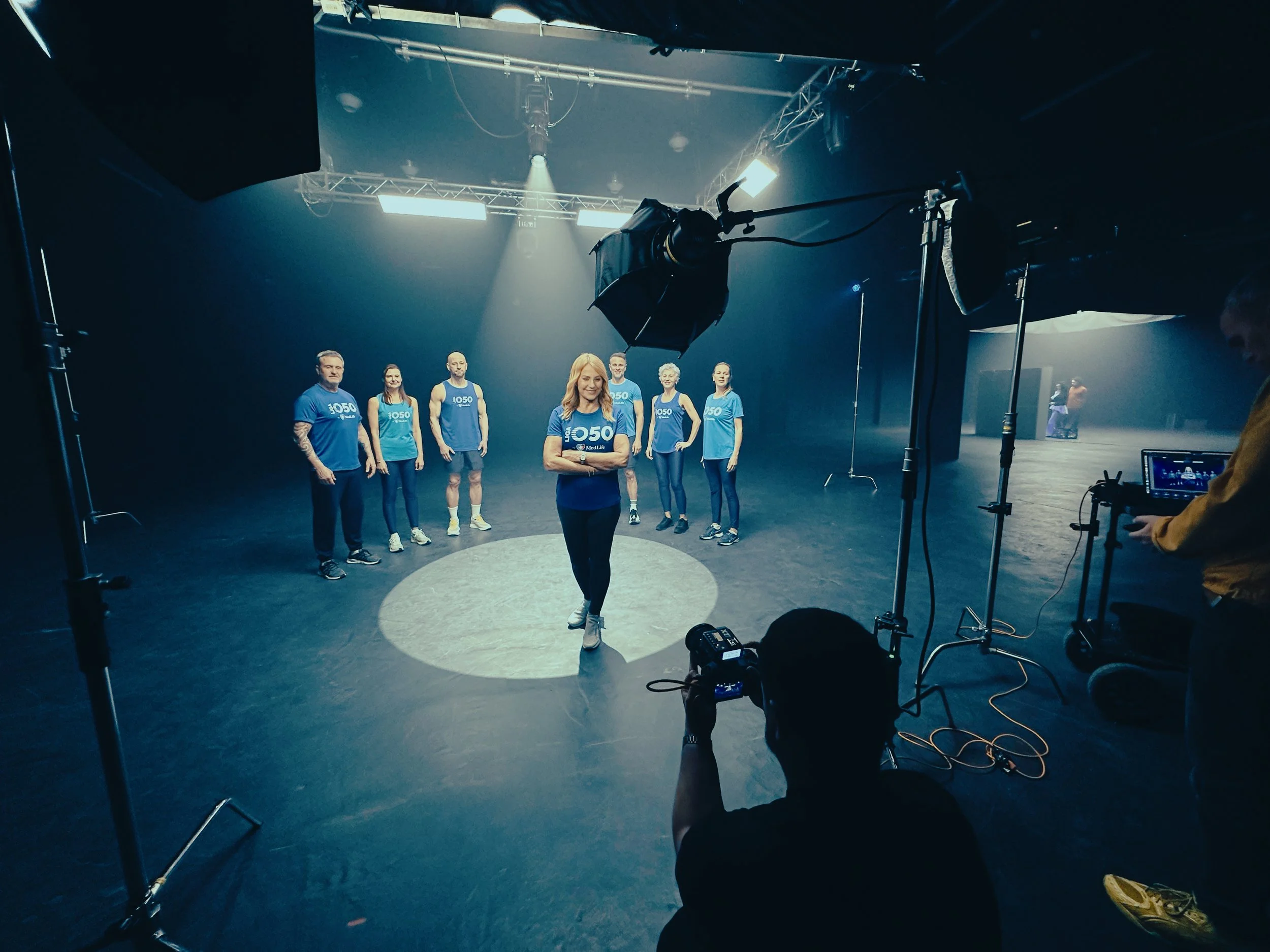 Group of people wearing blue T-shirts with a logo, standing on a stage or in a studio with professional lighting and equipment.