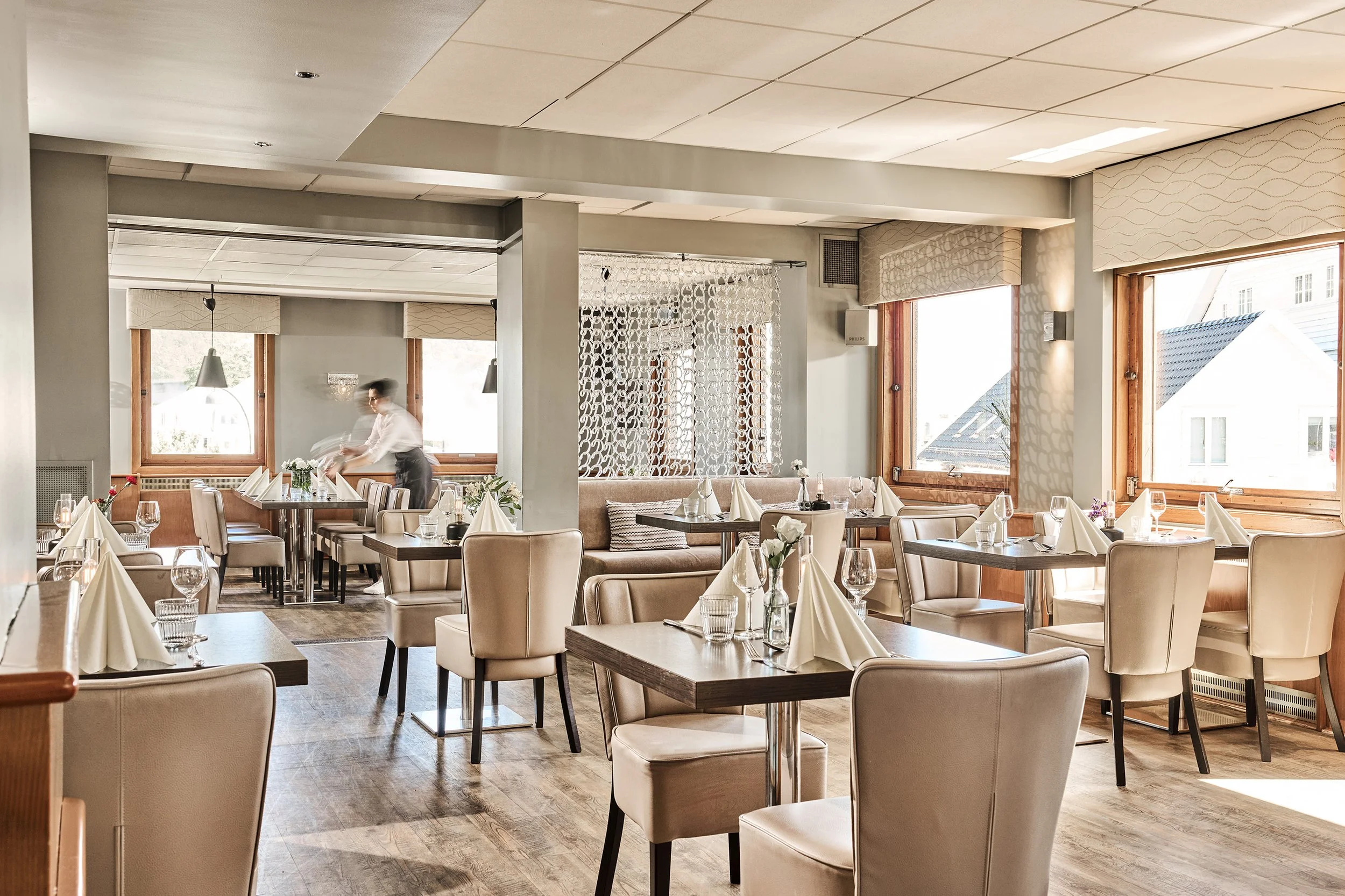 Innvendig av restaurant med bord dekket for gjester, store vinduer med utsikt, og en servitør som beveger seg.