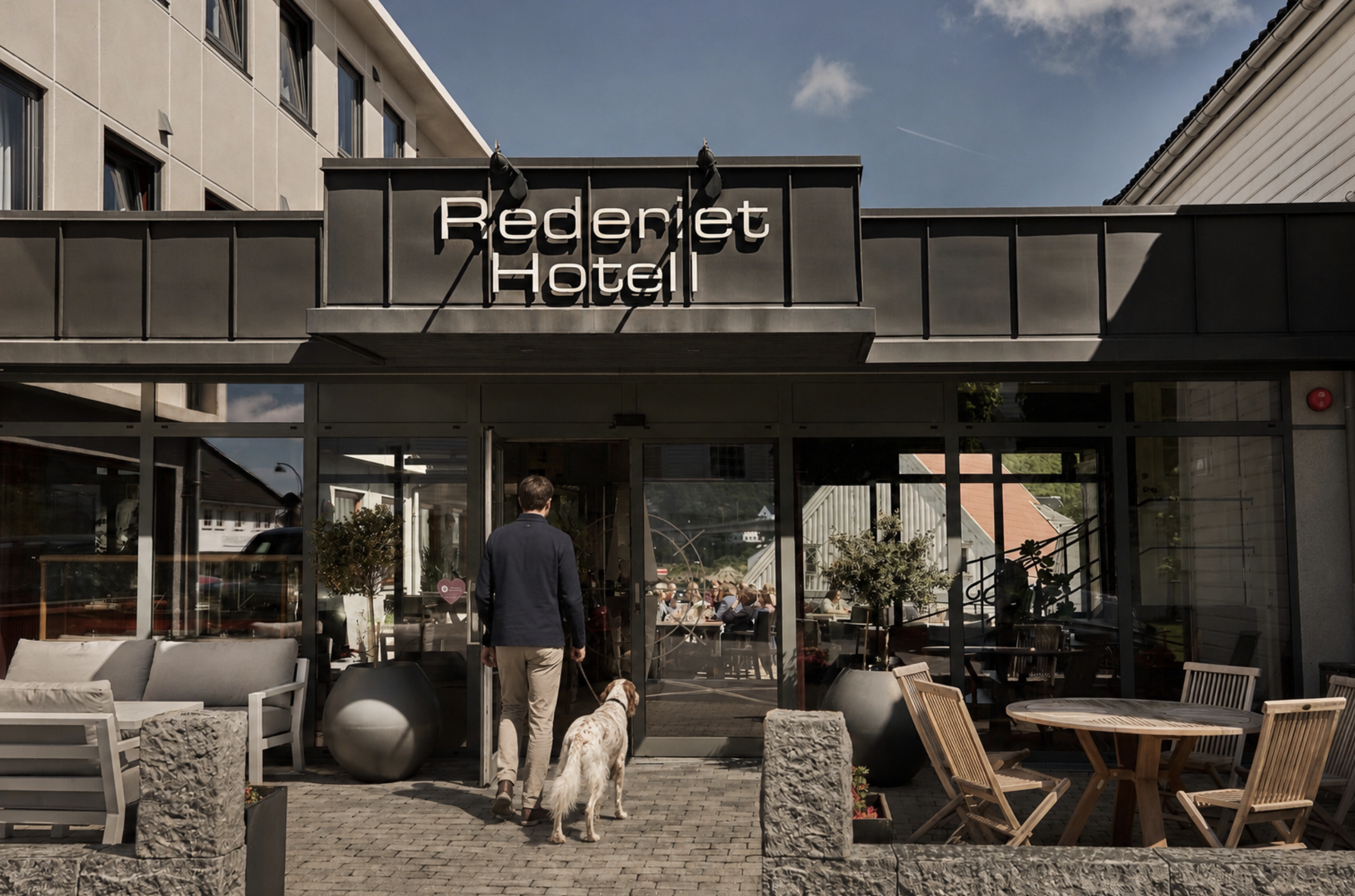 Utsiden av Redderiet Hotel med folk som spiser og koser seg utendørs. En person går inn i hotellet sammen med en hund. Utsiden har store glassvinduer, og det er sitteplasser med trebord og stoler.