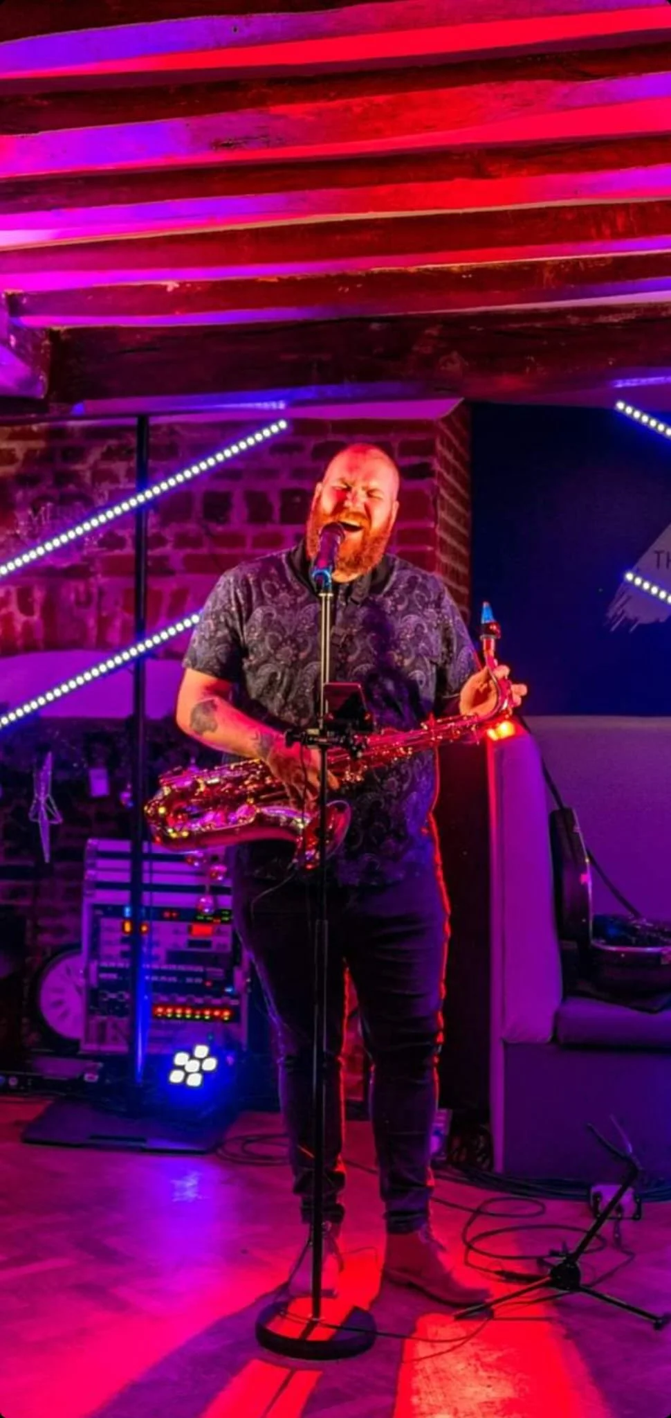 A male singer with a microphone and a saxophone performs on stage in a neon-lit venue with purple and pink lighting, exposed brick wall background, and electronic equipment nearby.