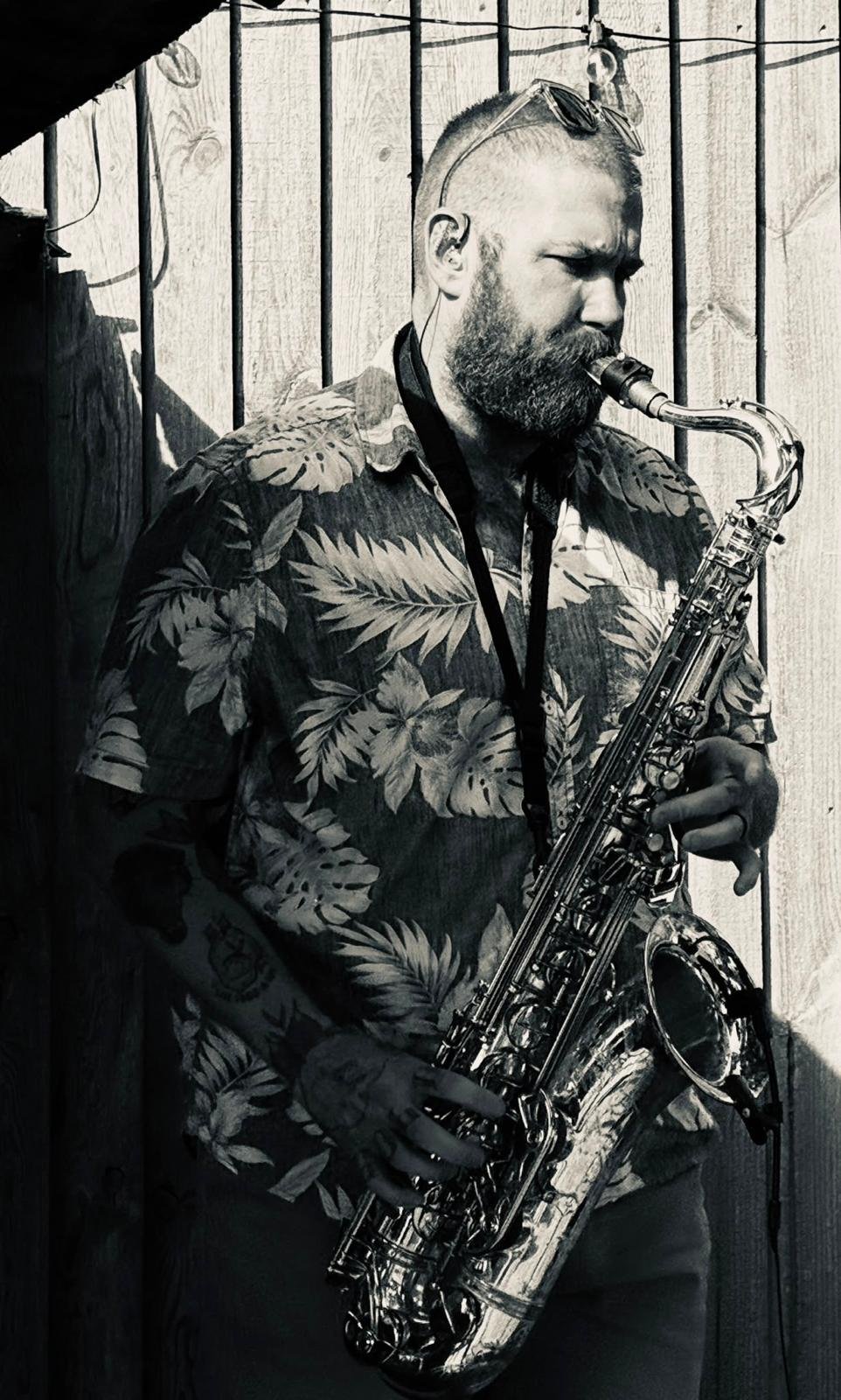 A man with a beard, sunglasses on his head, wearing a tropical print shirt, playing a saxophone against a wooden fence background.
