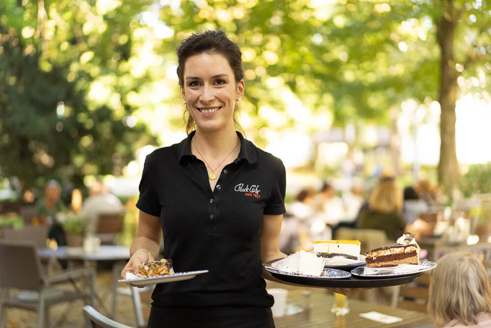 Eine Kellnerin im Freien, die ein Tablett mit verschiedenen Kuchenstücken trägt, umgeben von einem Garten mit Menschen im Hintergrund. Sie trägt ein schwarzes Poloshirt mit dem Aufdruck 'Park Café'. Der Hintergrund ist unscharf, mit grünen Bäumen und