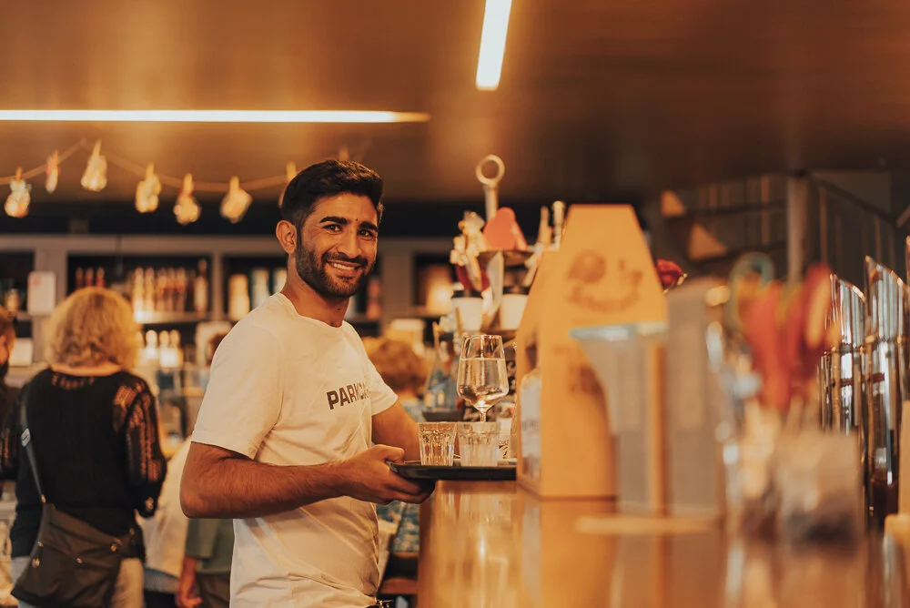 Barkeeper lächelt hinter einer Bar, hält ein Tablett mit Gläsern, im Hintergrund sind Flaschen und Gäste sichtbar.