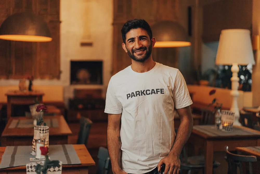 Ein lächelnder Mann in einem Café trägt ein weißes T-Shirt mit der Aufschrift "PARKCAFE". Im Hintergrund sind Tische, Stühle und warme Beleuchtung zu sehen.