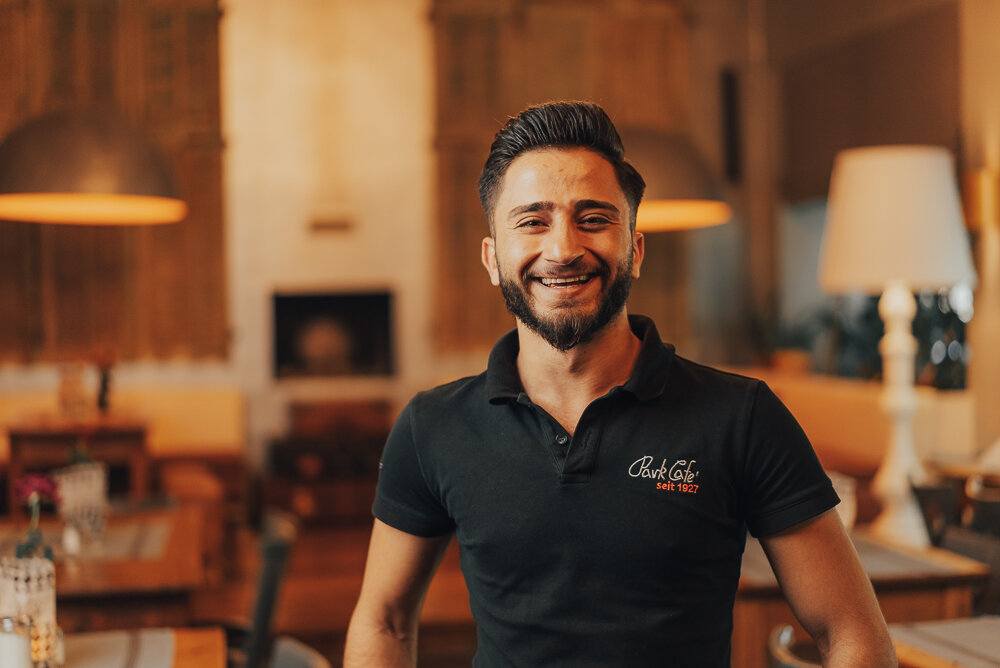 Mann in Café, lächelnd, trägt schwarzes Poloshirt mit "Park Cafe"-Logo, Innenansicht, warme Beleuchtung, Tische im Hintergrund.