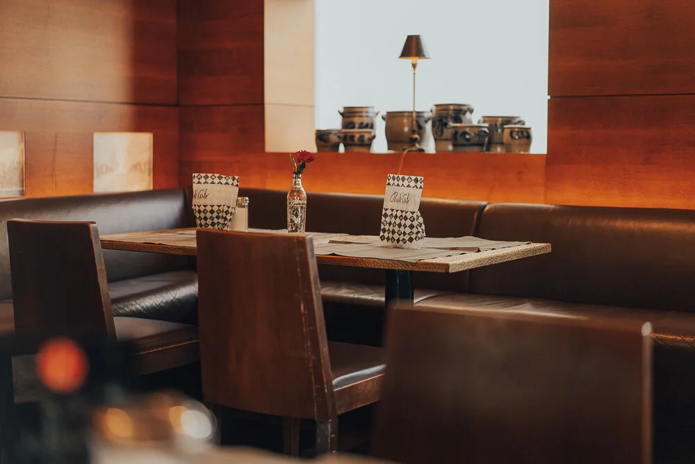 Innenaufnahme eines gemütlichen Restaurants mit Holzdekor, Lederbänken und Holztischen. Auf dem Tisch stehen eine Speisekarte und eine Vase mit Rose. Im Hintergrund sind dekorative Keramikvase zu sehen, neben einer kleinen Lampe für sanfte Beleuchtun