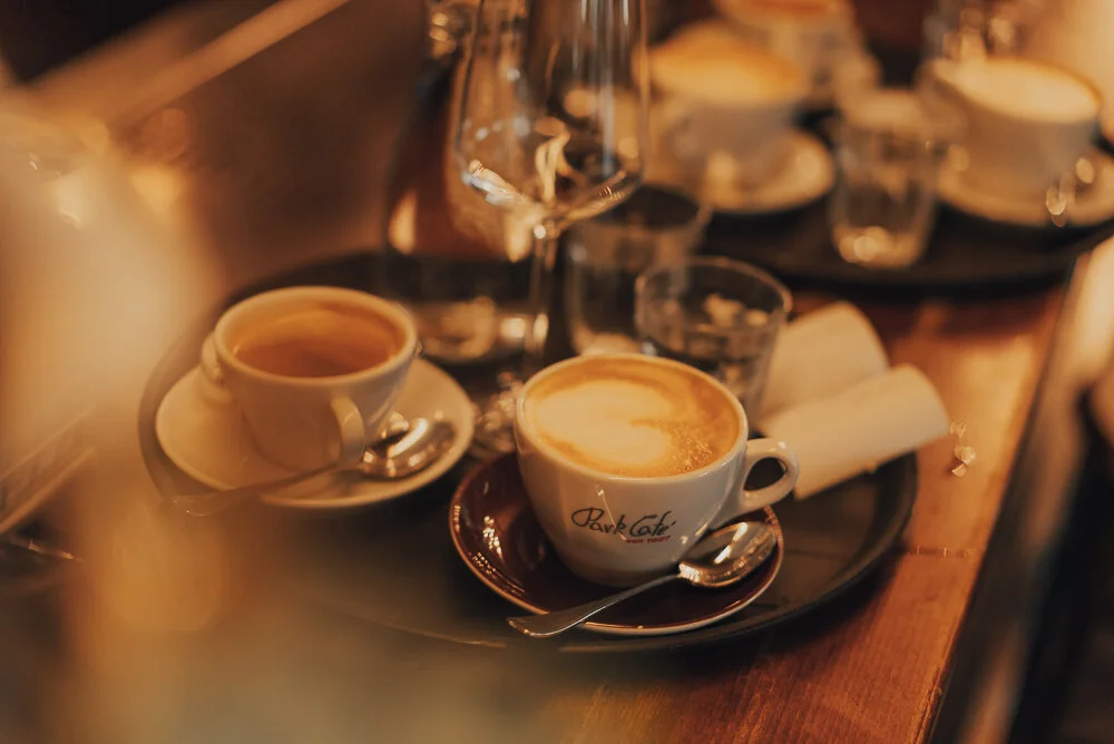 Tassen Kaffee auf einem Tisch in einem Café, umgeben von Glasflaschen und Gläsern. Die Atmosphäre ist warm und gemütlich, mit einem weichen Fokus.