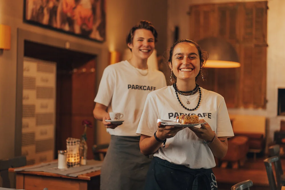 Zwei Café-Mitarbeiter in Parkcafé-T-Shirts lächeln, während sie Speisen und Getränke servieren. Im Hintergrund ist ein gemütlicher Innenraum mit Tischdekoration zu sehen.
