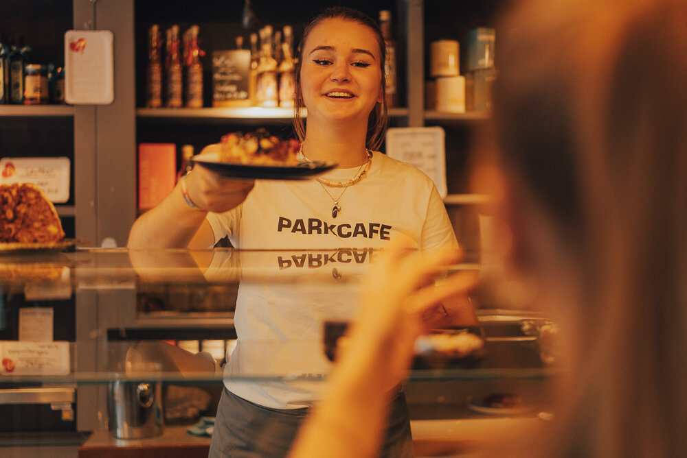 Frau hinter einer Theke in einem Café reicht einem Kunden einen Teller. Im Hintergrund sind Regale mit Flaschen und Verpackungen zu sehen. Die Frau trägt ein T-Shirt mit der Aufschrift 'PARKCAFE'.