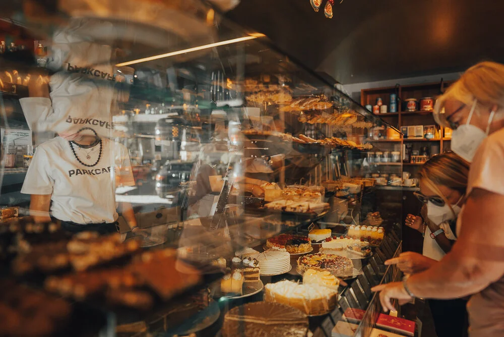 Drei Personen, zwei Erwachsene und ein Kind, betrachten ein Schaufenster voller Gebäck und Torten in einer Bäckerei. Eine Person hinter der Theke, andere tragen Schutzmasken.