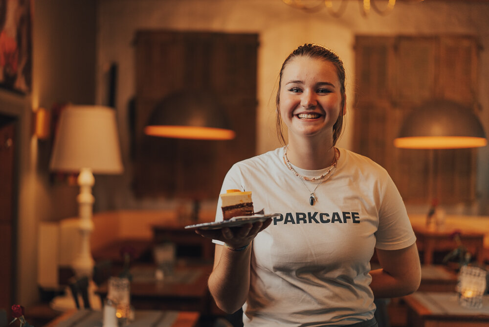 Junge Frau in einem Café, trägt ein weißes T-Shirt mit der Aufschrift 'PARKCAFE', hält einen Teller mit einem Kuchenstück. Im Hintergrund gemütliches Café-Ambiente mit Tischen und Lampen.