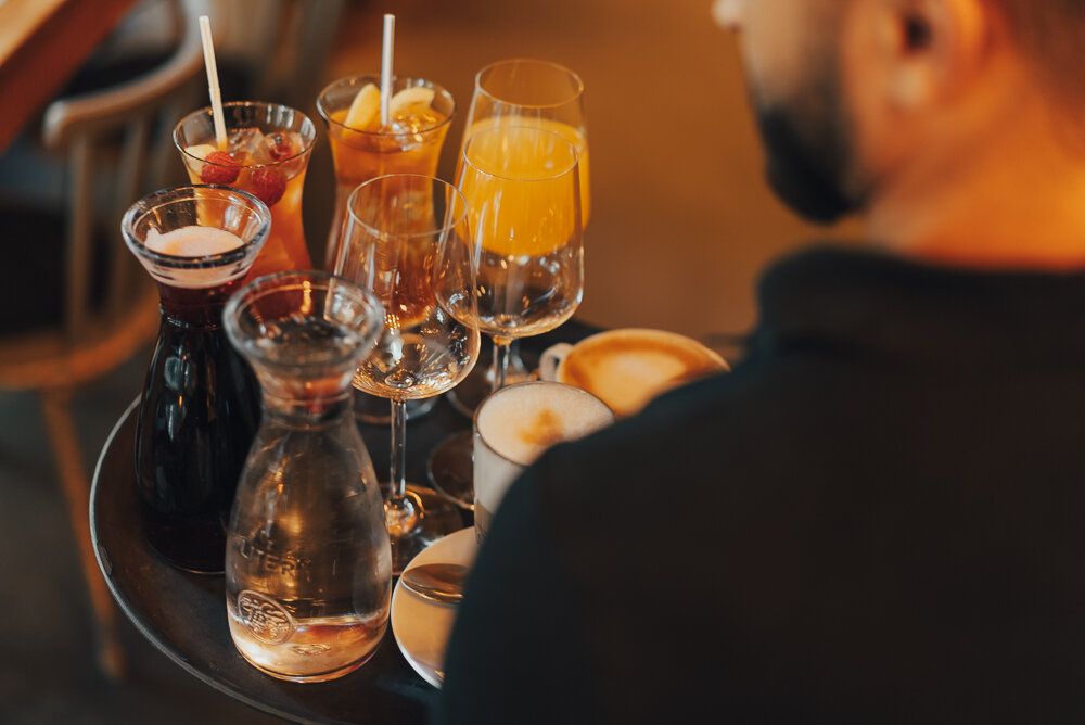 Nahaufnahme eines Tabletts mit verschiedenen Getränken, darunter Gläser mit Orangensaft, Cocktails, ein Krug mit klarer Flüssigkeit, ein Krug mit rotem Getränk und Tassen mit Kaffee. Eine Person hält das Tablett.