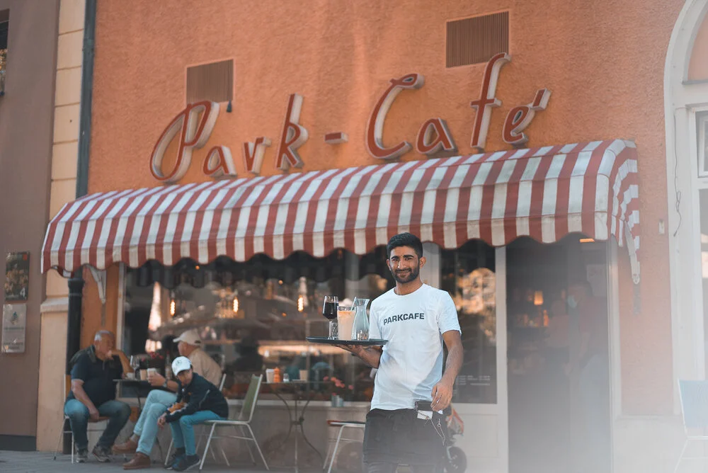 Ein Kellner trägt ein Tablett vor einem Café mit der Aufschrift "Park-Café". Das Café hat eine rot-weiße Markise. Im Hintergrund sitzen Gäste an einem Tisch.