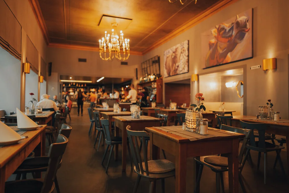 Ein stilvoll eingerichtetes Restaurant mit Holztischen, Stühlen und dekorativen Elementen wie Kerzen und Blumen auf den Tischen. Im Hintergrund ist ein Kronleuchter an der Decke und an den Wänden hängen farbige Gemälde. Es sind einige Menschen an den