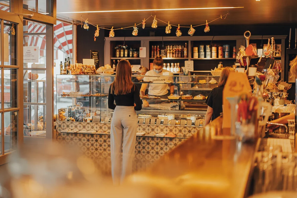 Innenansicht eines Cafés mit Kundschaft und Angestellten hinter einer Theke, die verschiedene Backwaren und Lebensmittel ausstellt.