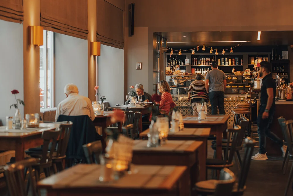 Innenansicht eines gemütlichen Cafés mit Holzmöbeln, gedeckten Tischen, mehreren Gästen an Tischen und einer Theke mit Baristas im Hintergrund.