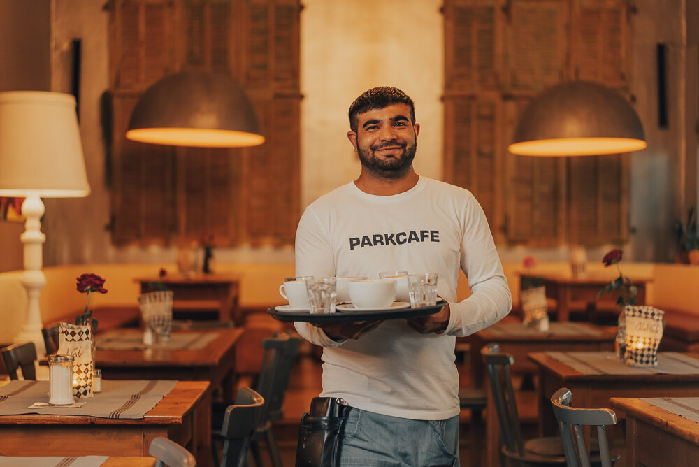 Ein lächelnder Mann in einem Café mit "PARKCAFE" auf seinem weißen Shirt hält ein Tablett mit Tassen. Der Raum ist gemütlich mit Holzmöbeln und warmem Licht dekoriert.