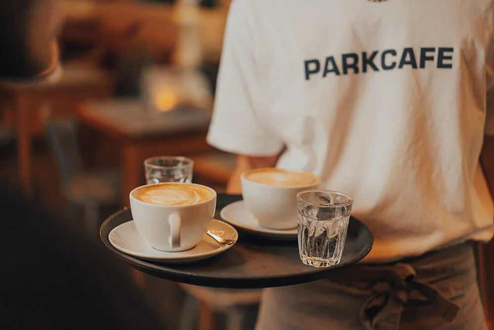 Ein Kellner oder eine Kellnerin trägt ein Tablett mit zwei Tassen Kaffee und zwei Gläsern Wasser. Auf dem T-Shirt steht 'PARKCAFE'. Der Hintergrund zeigt ein Café-Interieur.