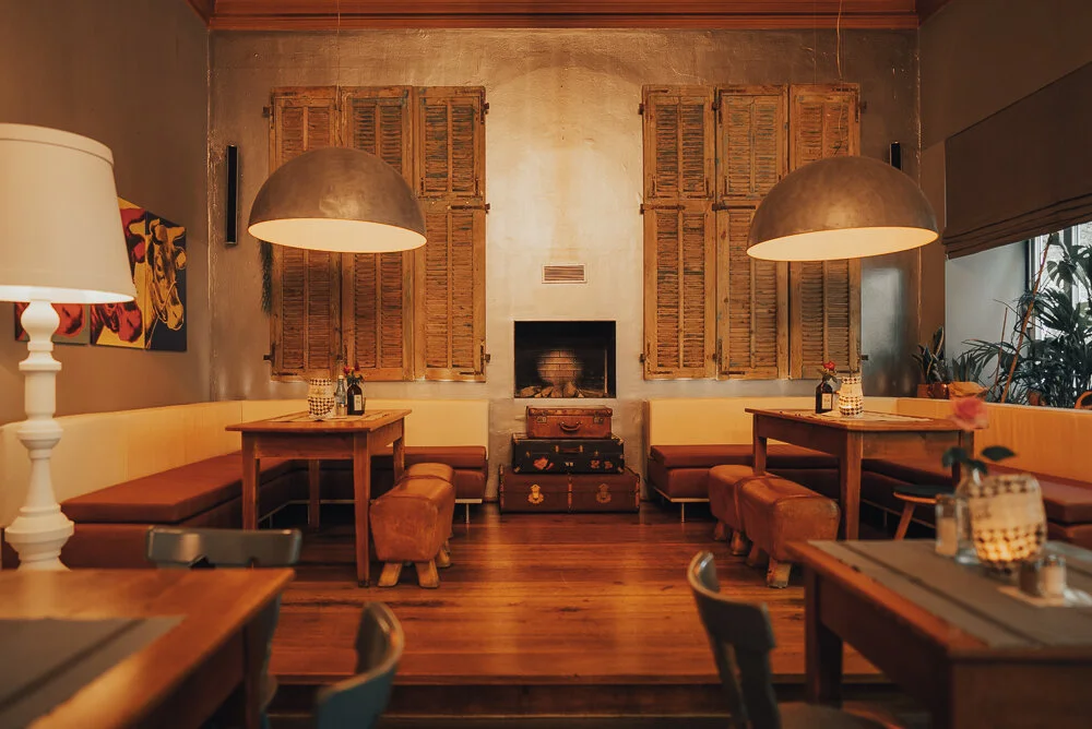 Innenansicht eines gemütlichen Restaurants mit Holzmöbeln, zwei großen Hängelampen, Pflanzen, einer Wand mit Jalousien, Holztischen und gepolsterten Bänken. Warme Beleuchtung und dekorative Elemente sorgen für ein einladendes Ambiente.