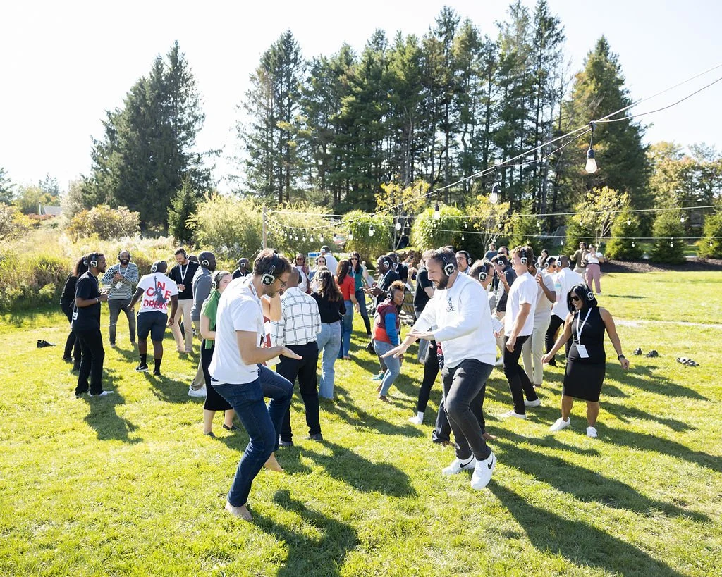 People dancing outdoors at a sunny park with trees and string lights overhead.