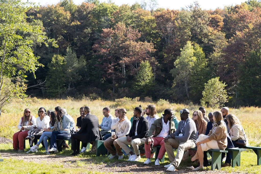 A group of people sitting on benches outdoors in a grassy field surrounded by trees with autumn foliage.