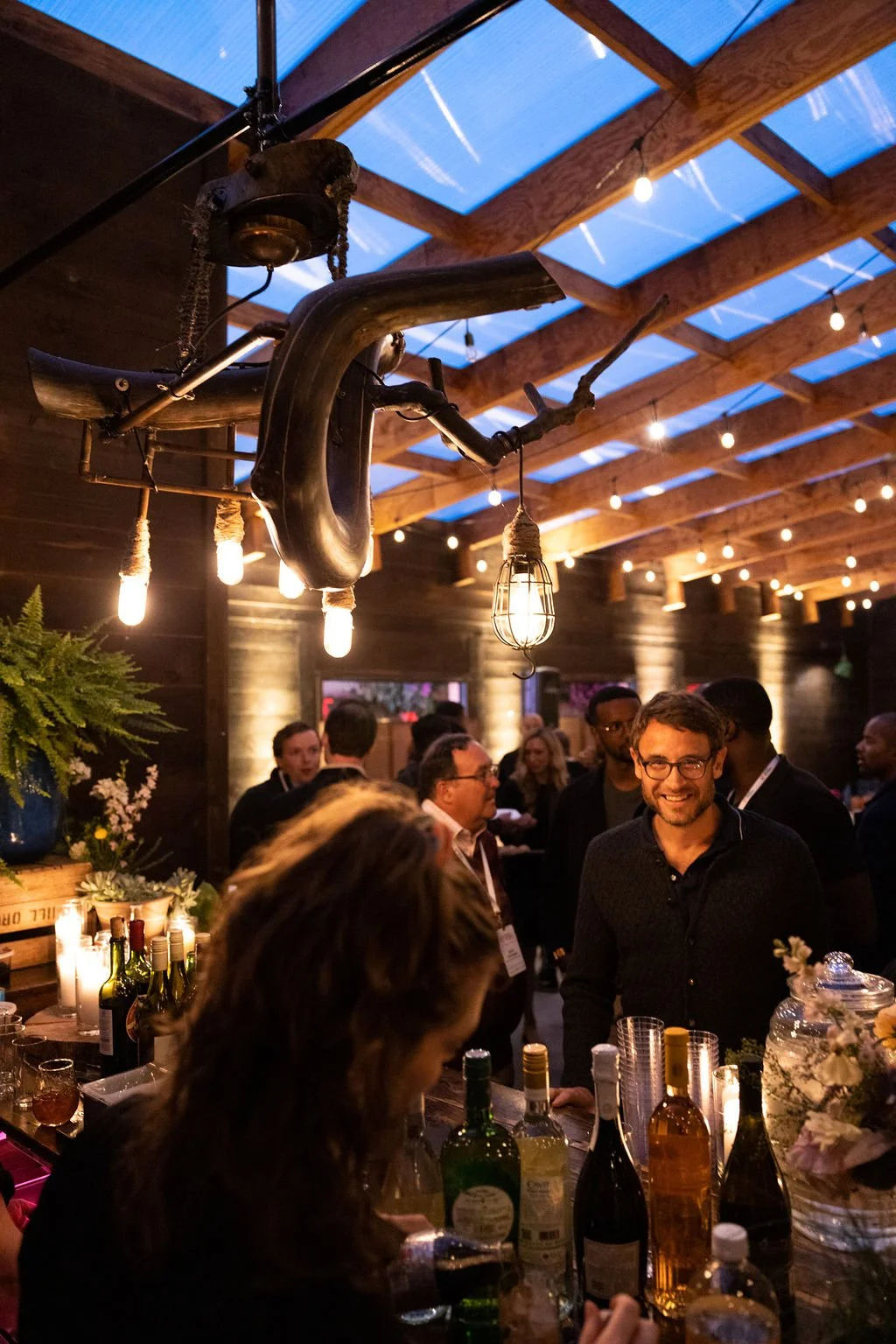 A social gathering at a bar or restaurant with a rustic wooden ceiling and string lights. There are multiple people socializing, a smiling man wearing glasses, bottles of alcohol on the bar, and a unique hanging light fixture made from car parts.