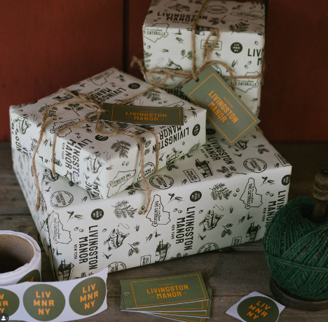 Wrapped gift boxes with Livingston Manor themed wrapping paper and tags on a rustic wooden surface, next to a spool of green thread.