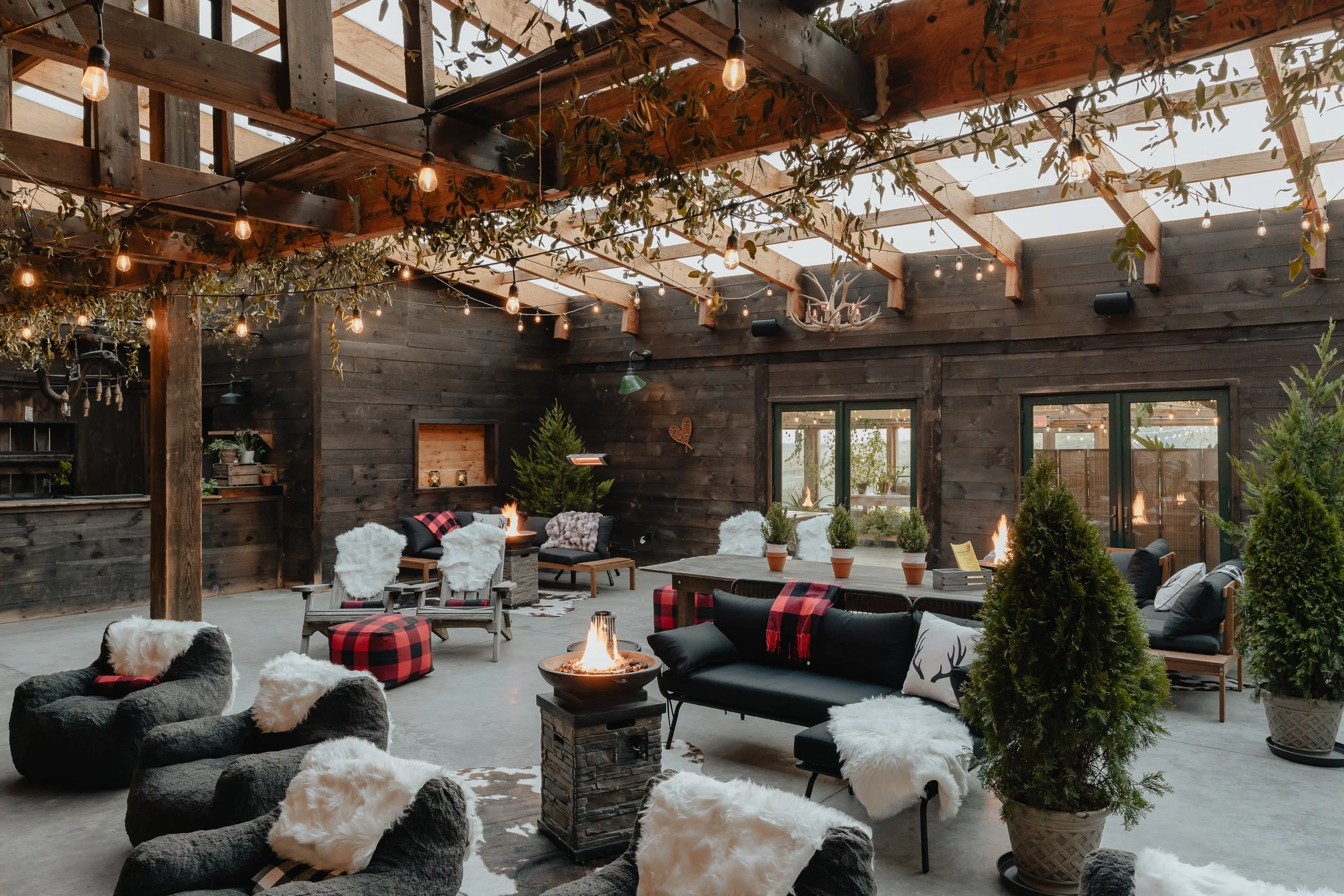 Indoor rustic patio with wooden walls and ceiling, string lights, potted plants, cozy seating with fur throws, and fire pits creating a warm, inviting atmosphere.