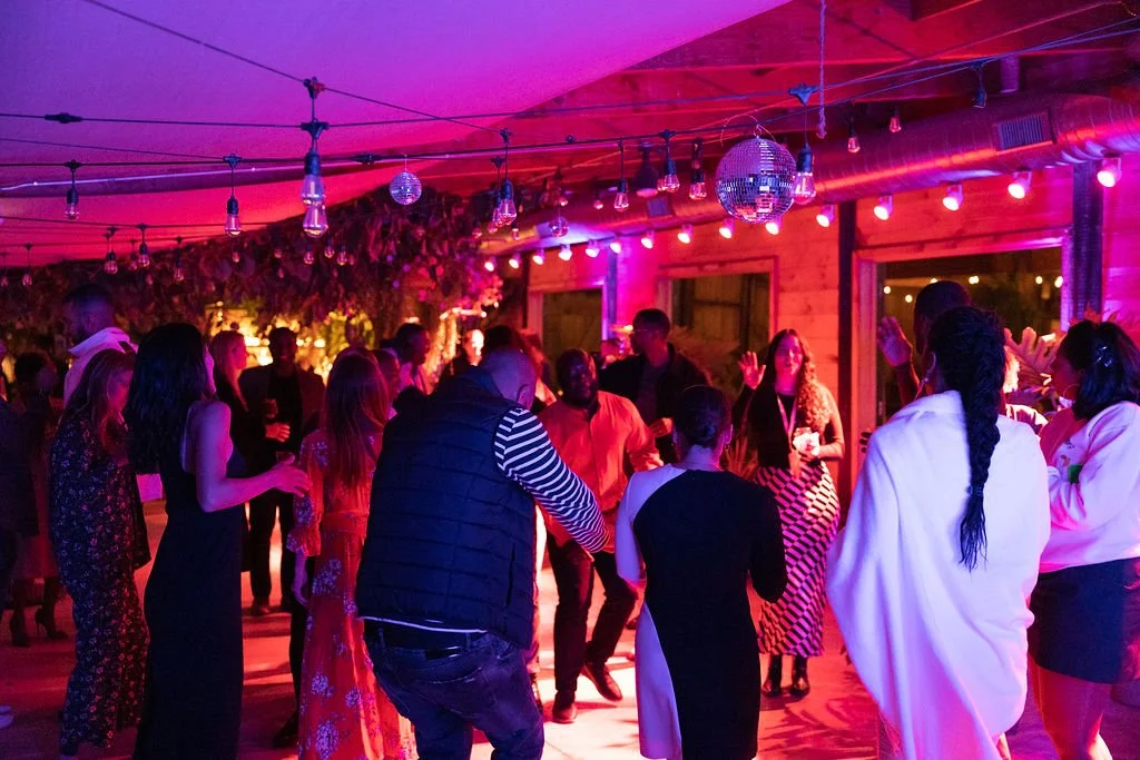 People dancing and socializing at a lively indoor party with colorful pink and purple lighting, disco balls, and string lights hanging from the ceiling