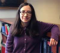 Woman wearing a purple cardigan with shoulder length brown hair and glasses leaning against book cases