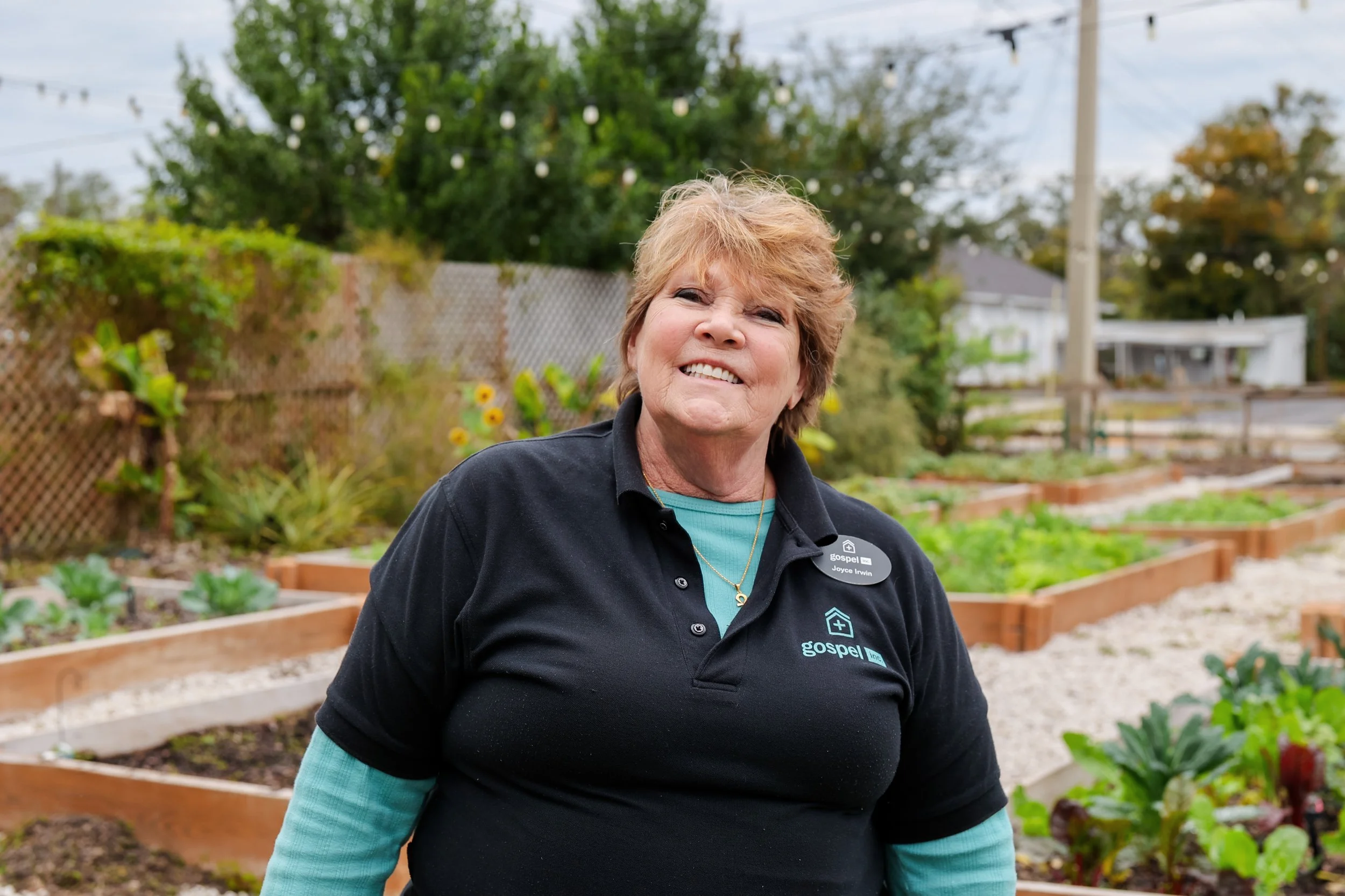 A woman smiling in a garden with raised beds and plants. She is wearing a black polo with the logo 'gospel' and a name tag.