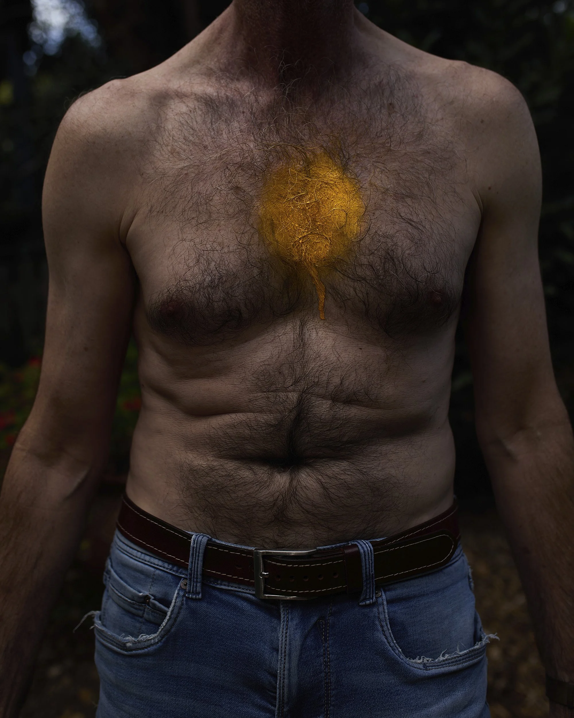 A cropped photograph of a man wearing jeans with a bare torso and a spray painted yellow spot on his chest.