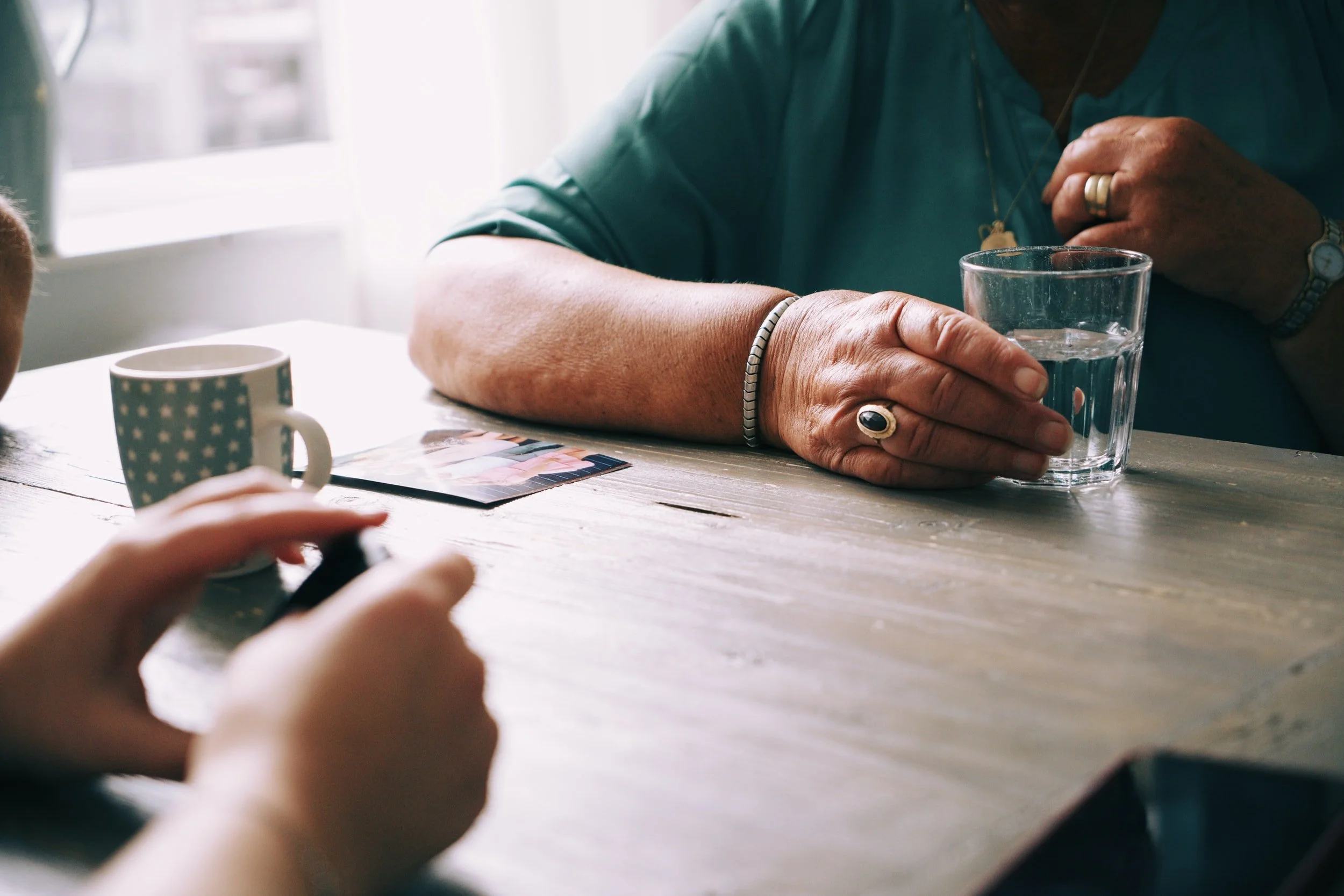 Close-up of an elderly person's hand with rings, holding a glass of water on a wooden table, with another person's hand and a mug with a star pattern nearby.