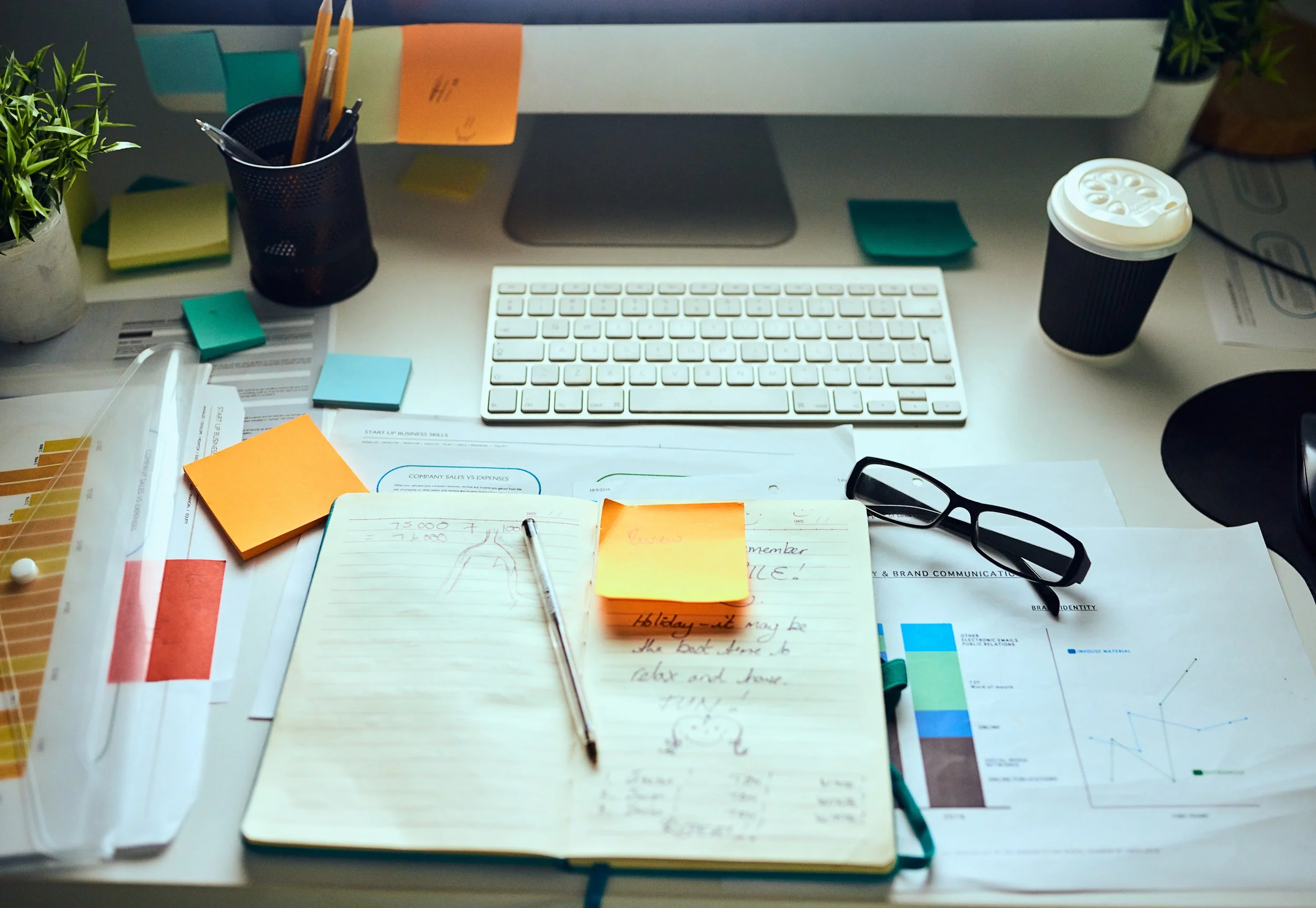 Desk cluttered with papers, sticky notes, glasses, a keyboard, a plant, a coffee cup, and stationery.