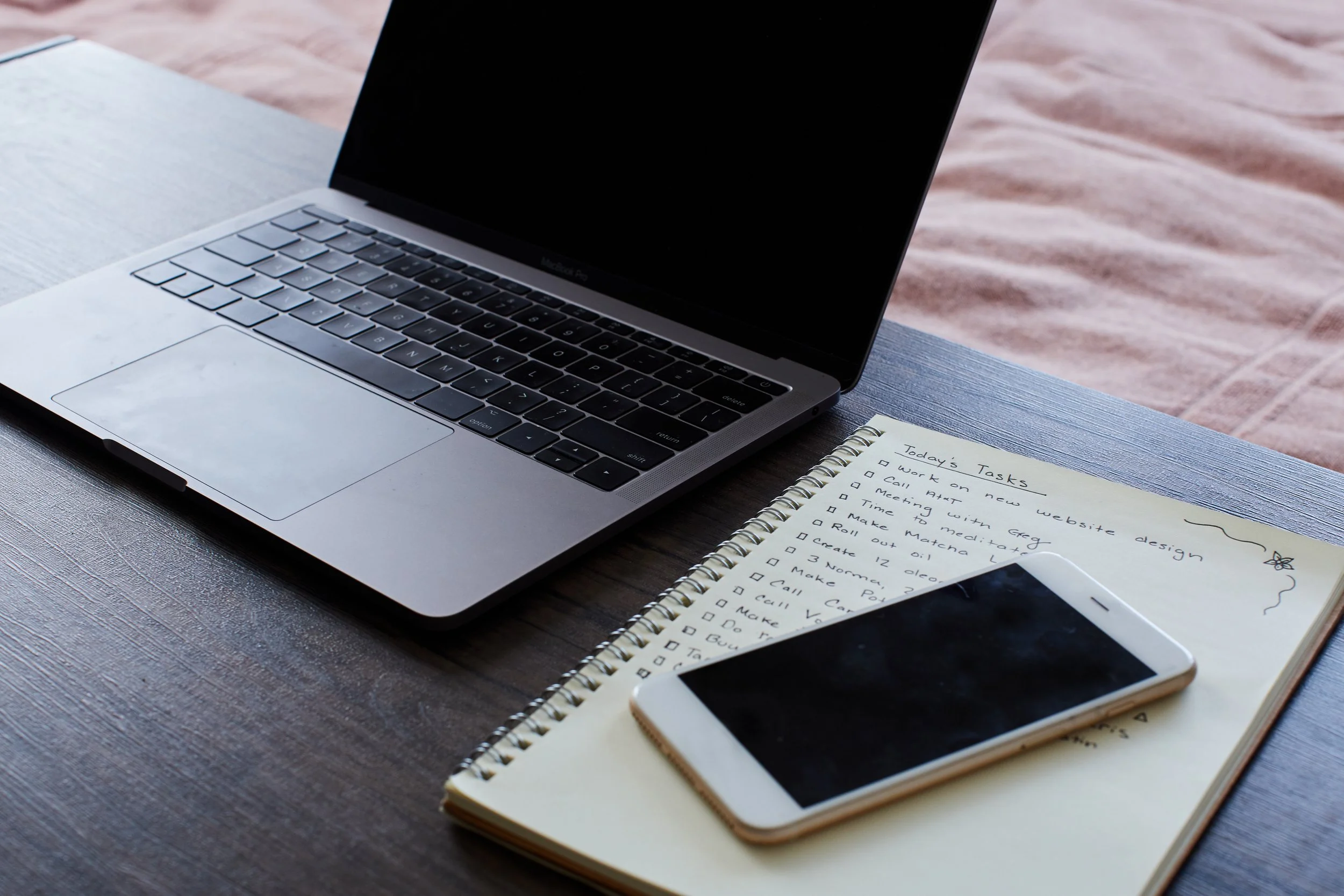 Open MacBook laptop on wooden desk next to a handwritten to-do list in a spiral notebook and a white smartphone.
