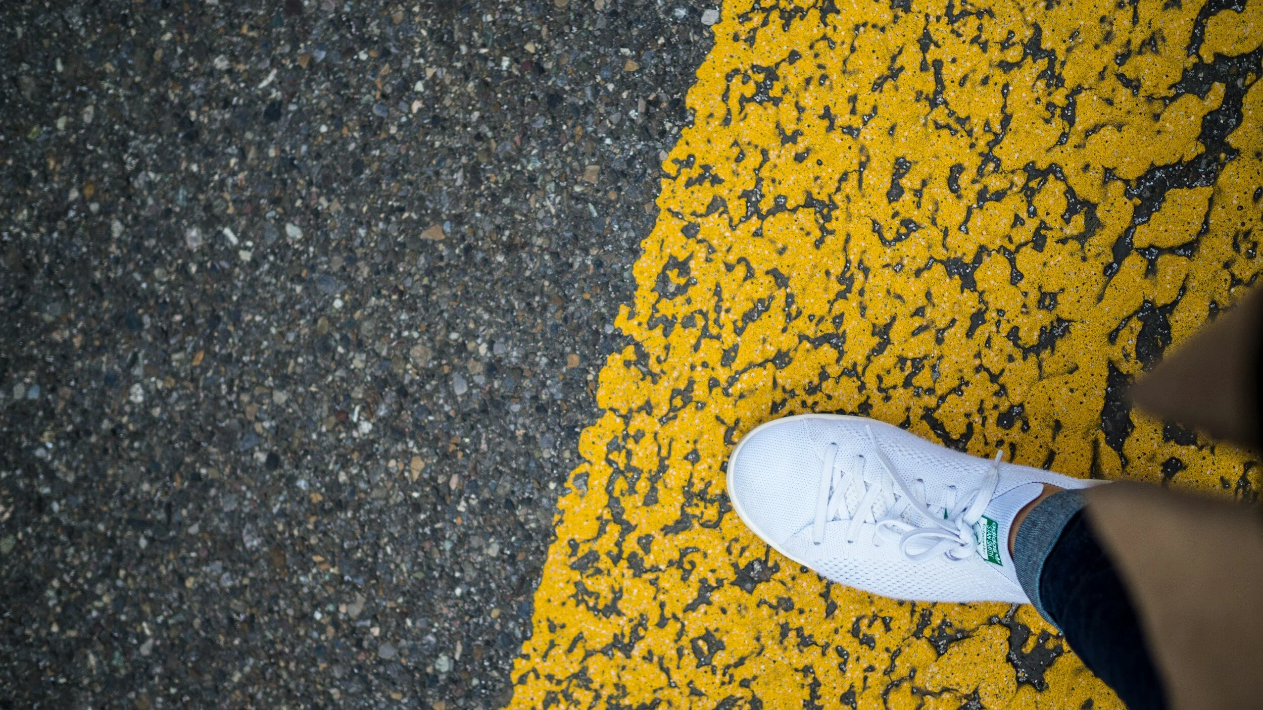 A person's foot in a white sneaker standing on a painted yellow line on a rough asphalt road.