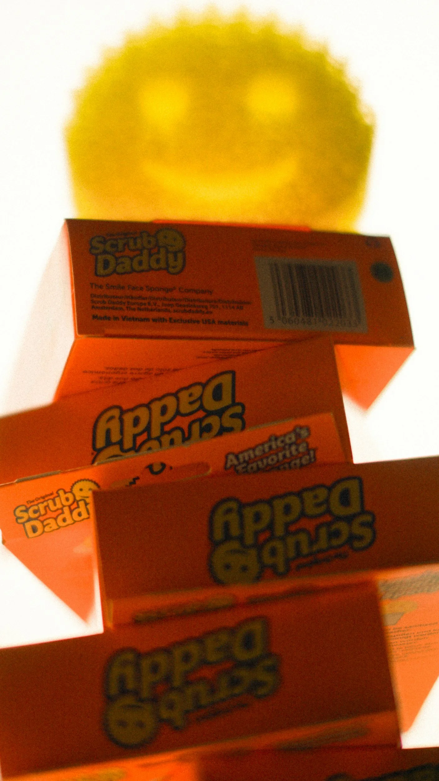 A close-up of orange boxes of Scrub Daddy, a smiling face sponge, stacked in a row. The boxes feature branding and product information.