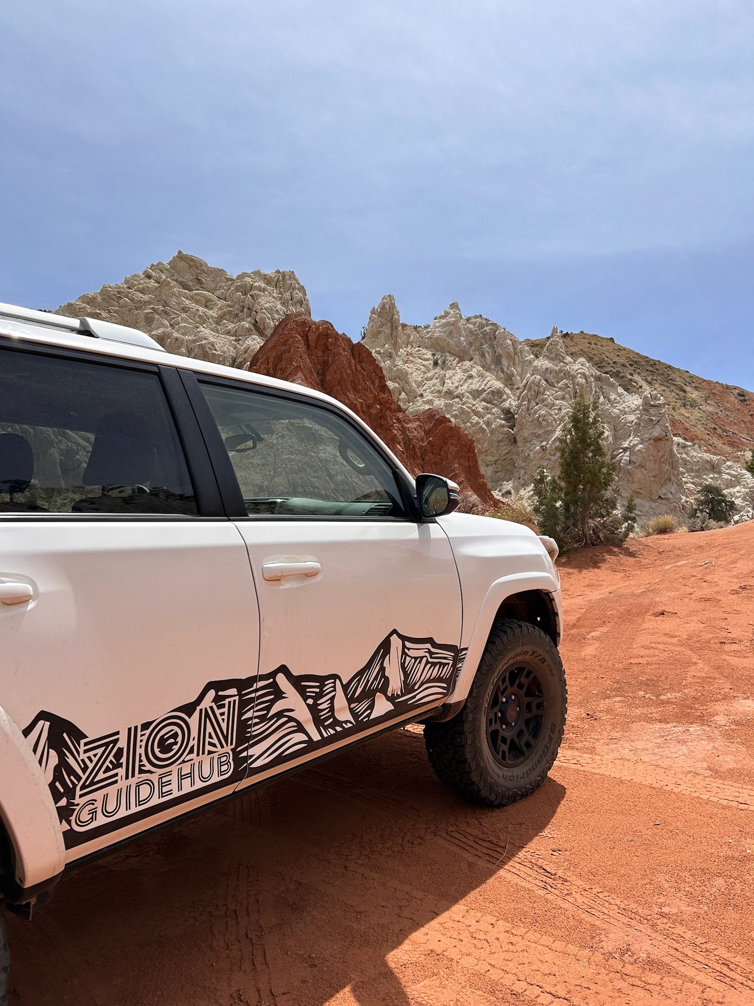 A white vehicle with Arizona Guide Hub logo parked on a dirt trail in a desert landscape, with red and white rocky mountains and sparse vegetation in the background under a partly cloudy sky.