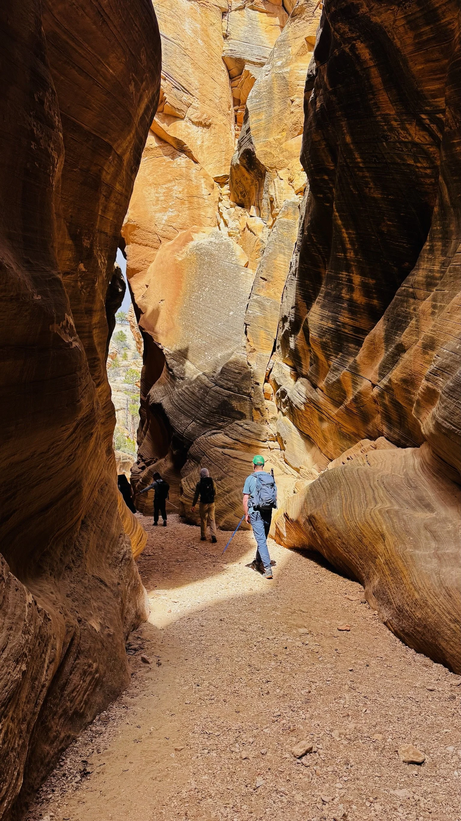 Hikers walking through a narrow slot canyon with towering, layered sandstone walls in desert terrain.