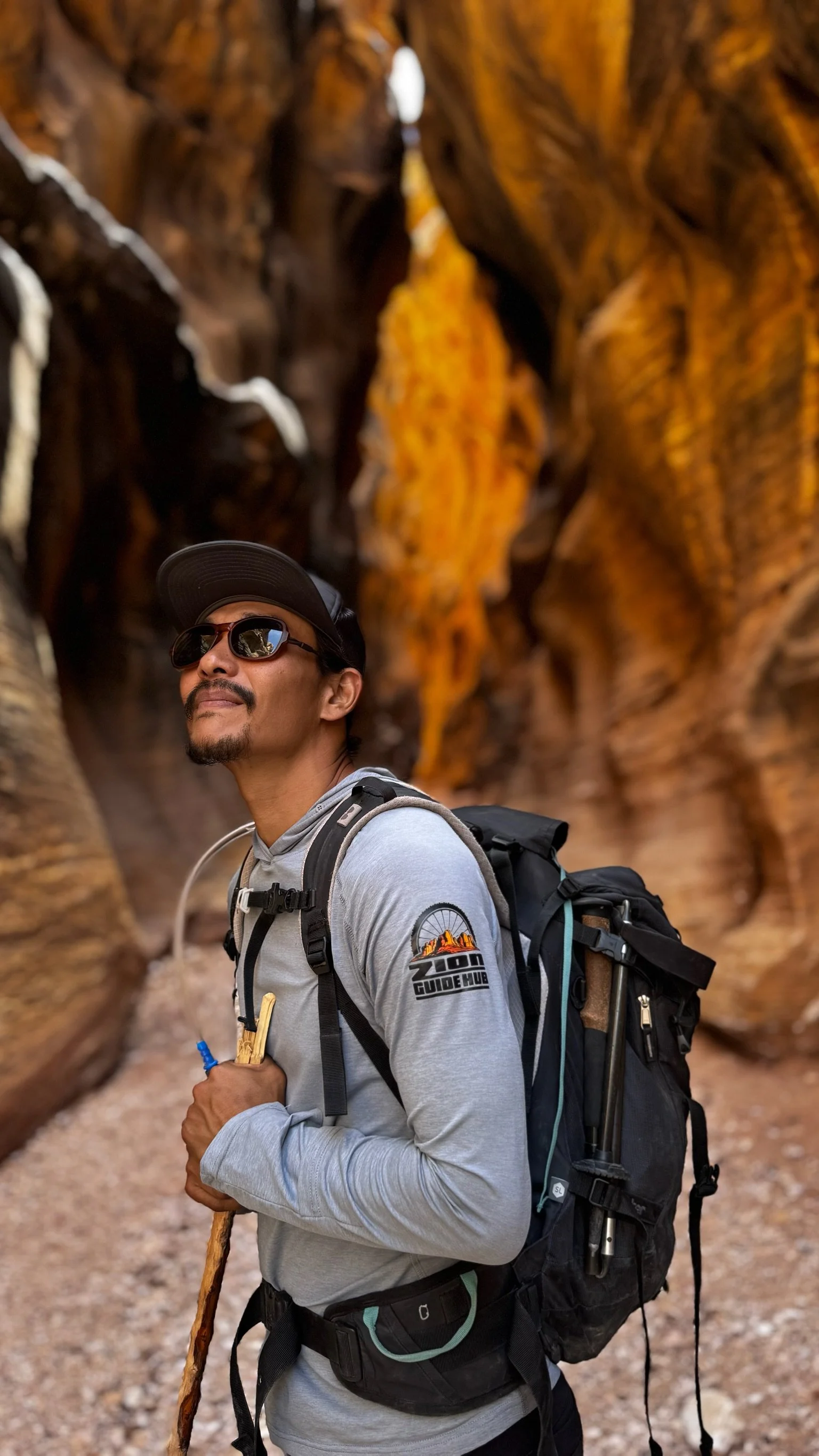 A man wearing sunglasses and a cap hiking through a narrow canyon with reddish-brown rock walls, carrying a large backpack and a wooden stick.