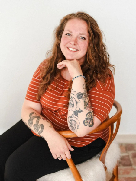 Smiling woman with red hair, tattoos on her arms, wearing a red and white striped shirt, sitting on a wooden chair against a plain white wall.