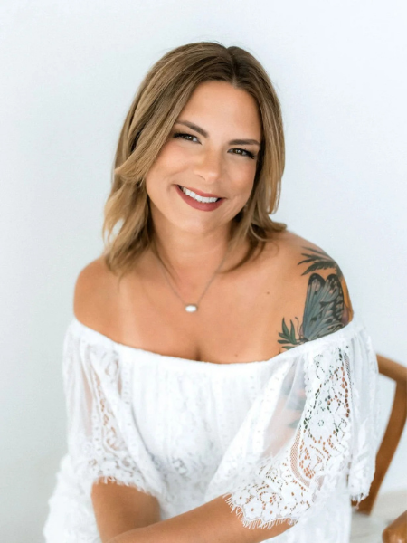 A young woman with shoulder-length light brown hair and a tattoo on her right shoulder smiling, wearing an off-the-shoulder white lace top and a delicate necklace, sitting against a light background.