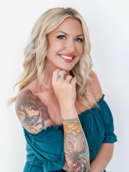 A smiling blonde woman with tattoos on her left arm, wearing an off-the-shoulder blue top, and holding her chin with her right hand, against a white background.