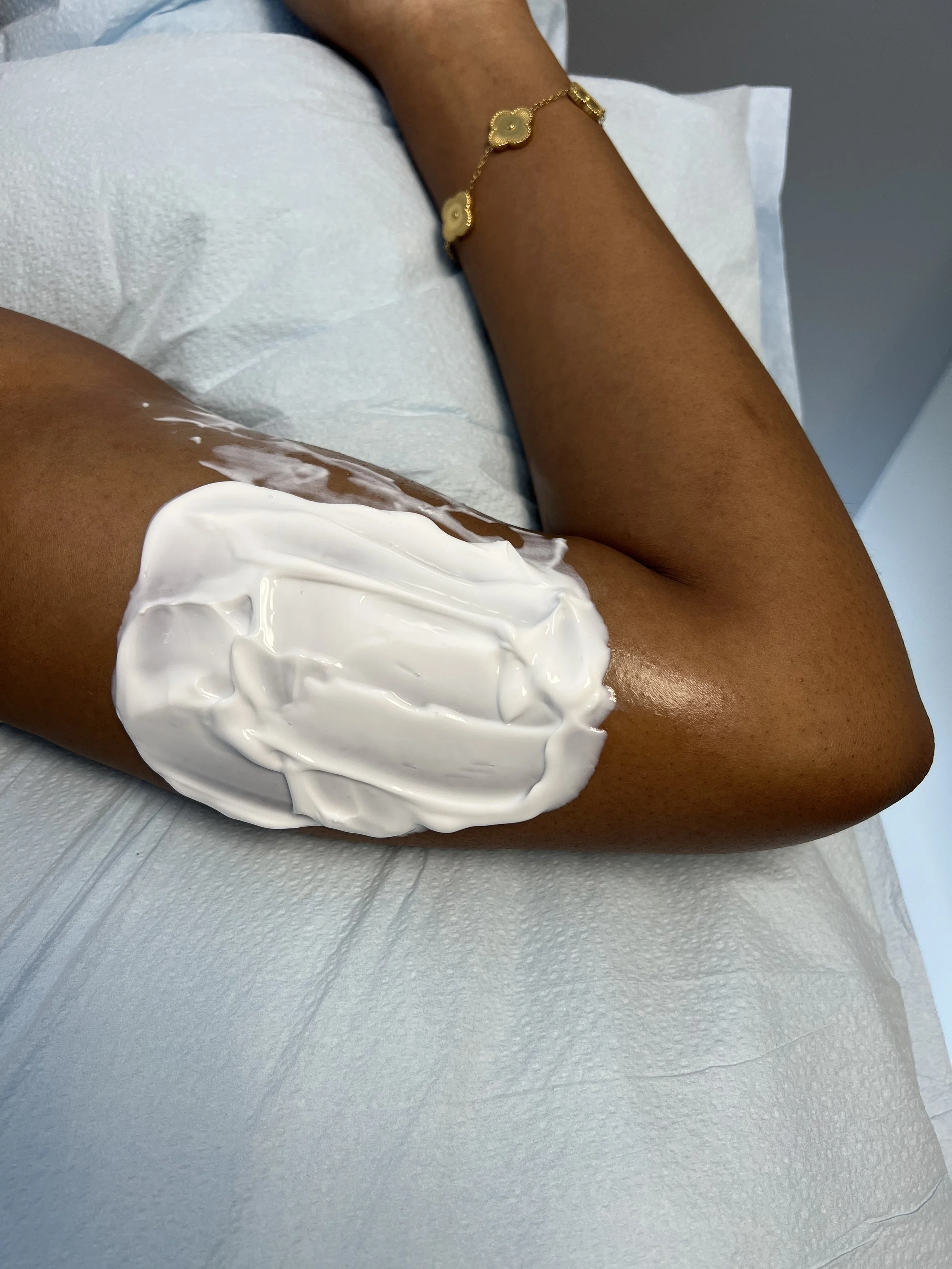 A tube of acrylic paint labeled "Gouache" lying on a white surface, with a blurred bottle of paint in the background and a piece of artwork with white flowers on the left.