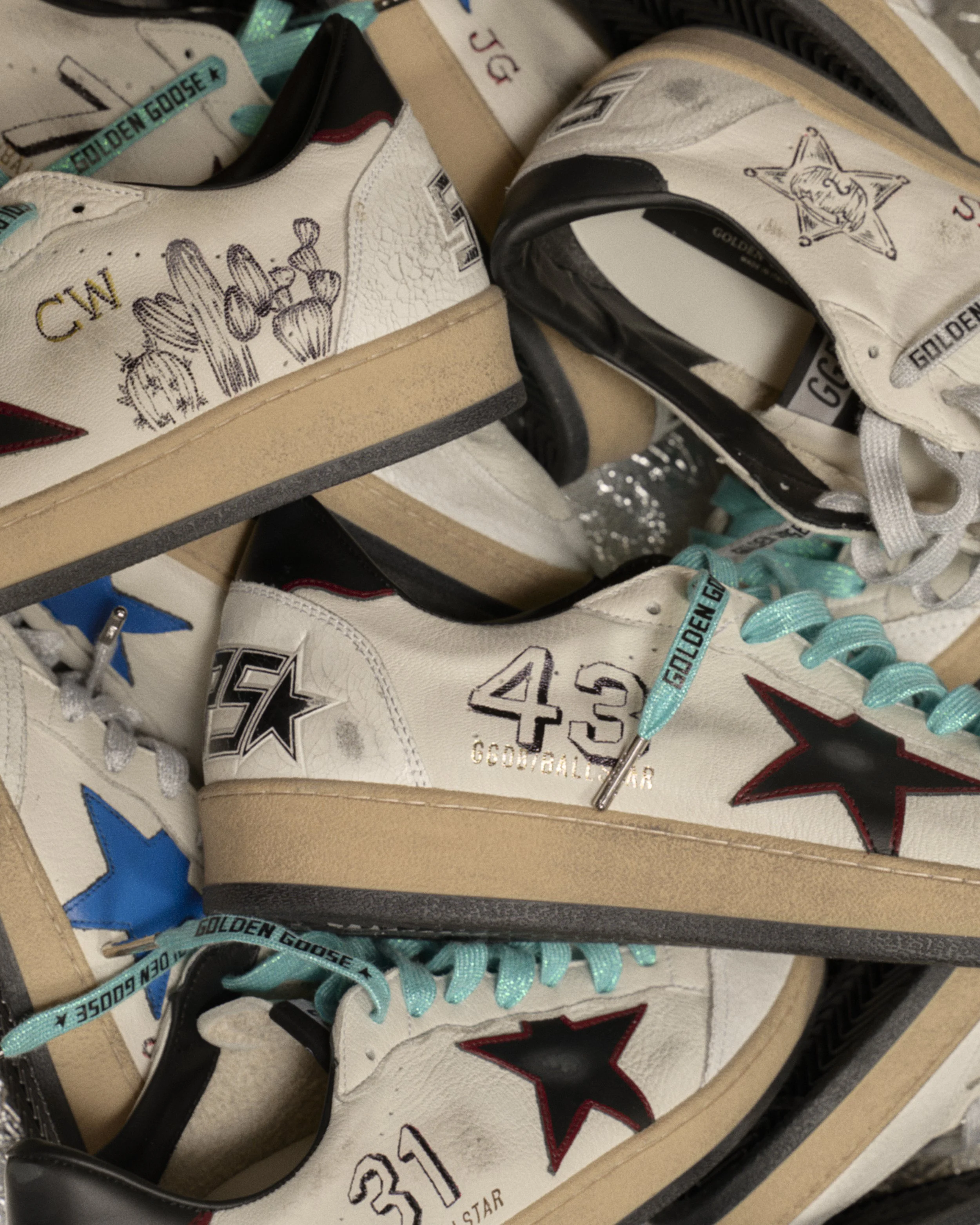 Multiple pairs of white Golden Goose sneakers with black and red accents, each decorated with hand-drawn illustrations and text, displayed in a shoebox.