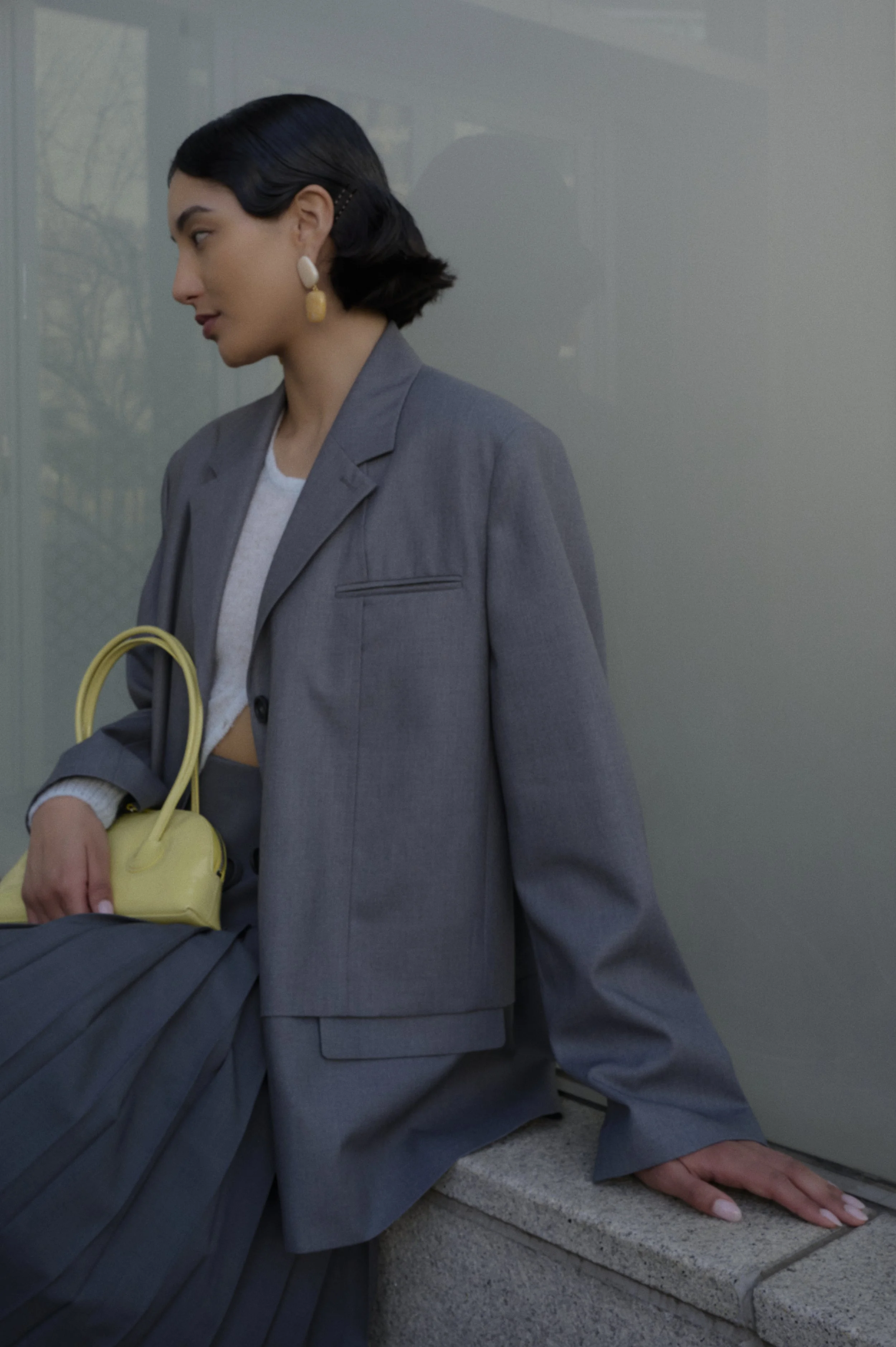 A woman with short black hair, wearing a gray suit and large earrings, sitting on a stone ledge outside. She holds a yellow purse and a gray garment or bag in her lap. She has manicured nails and is glancing to her left, with her right hand resting o